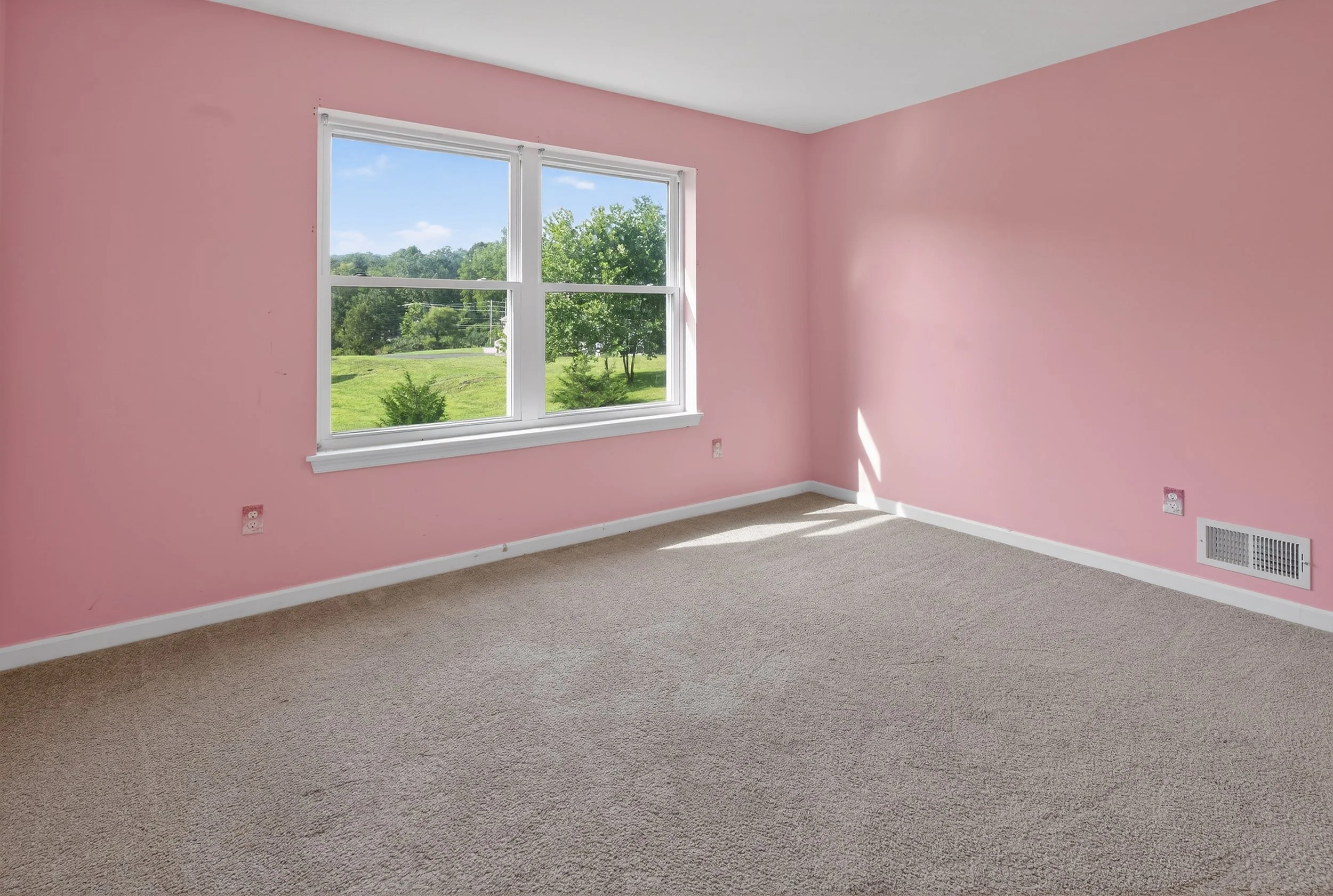 Empty room with pink walls, beige carpet, large window with a view of green trees and grass, white baseboards, and a floor vent.