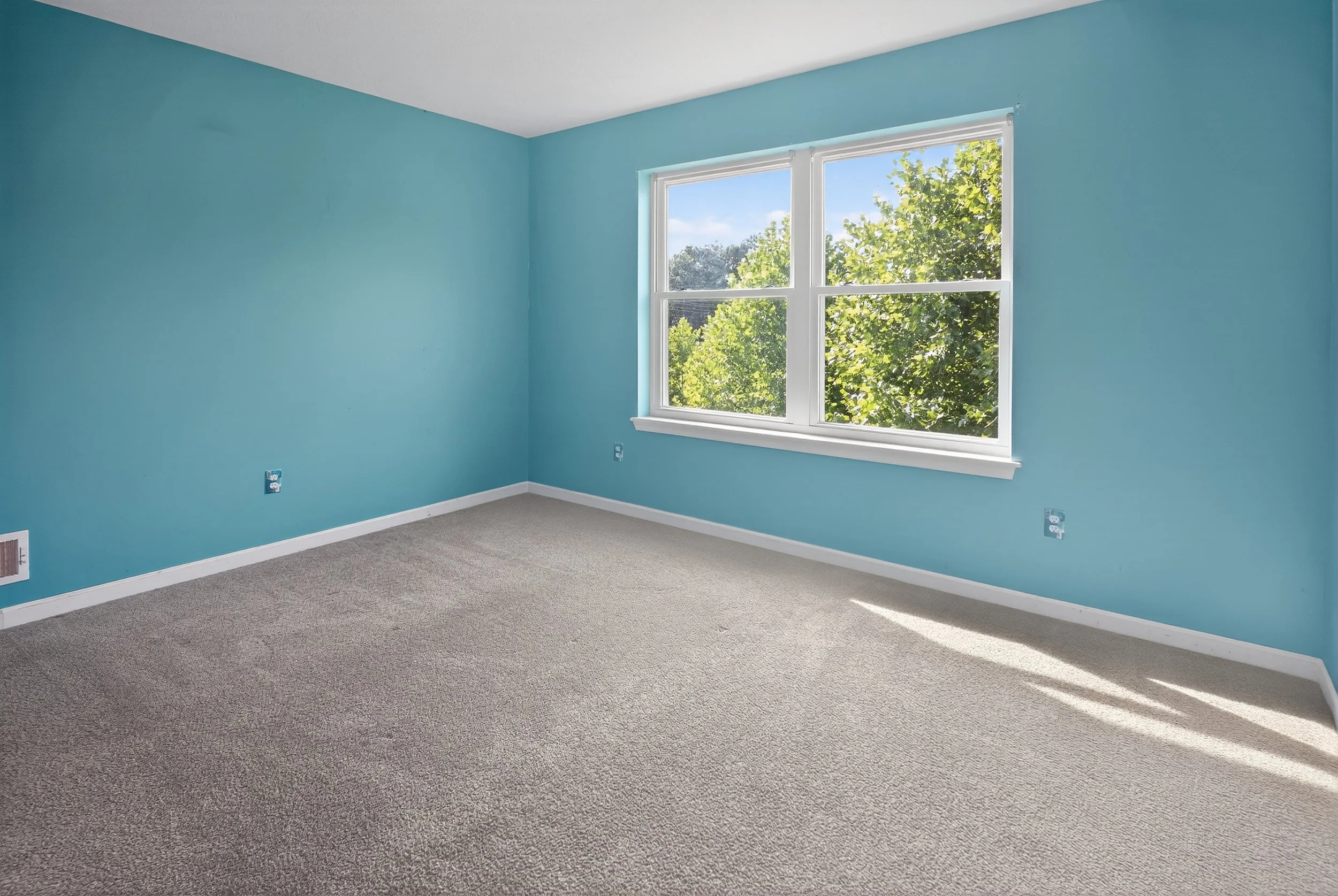 Empty room with turquoise walls, a large window showing green trees outside, beige carpeted floor, and white trim, no furniture.