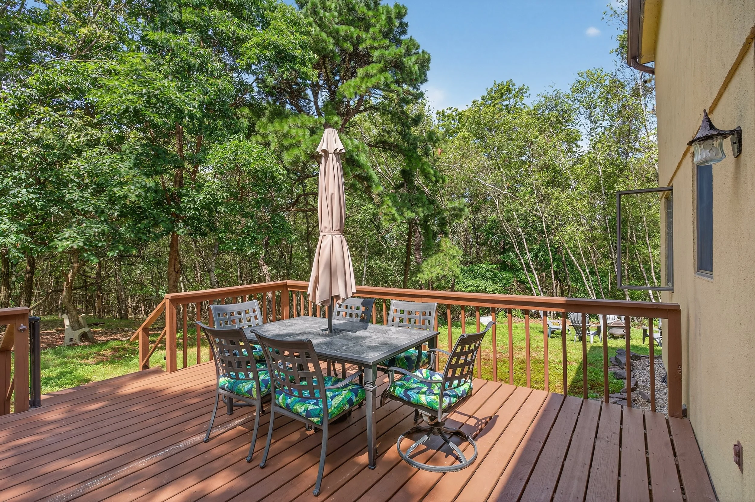 Backyard deck with a table, six chairs with colorful cushions, and a closed umbrella, surrounded by trees and greenery under a clear blue sky.