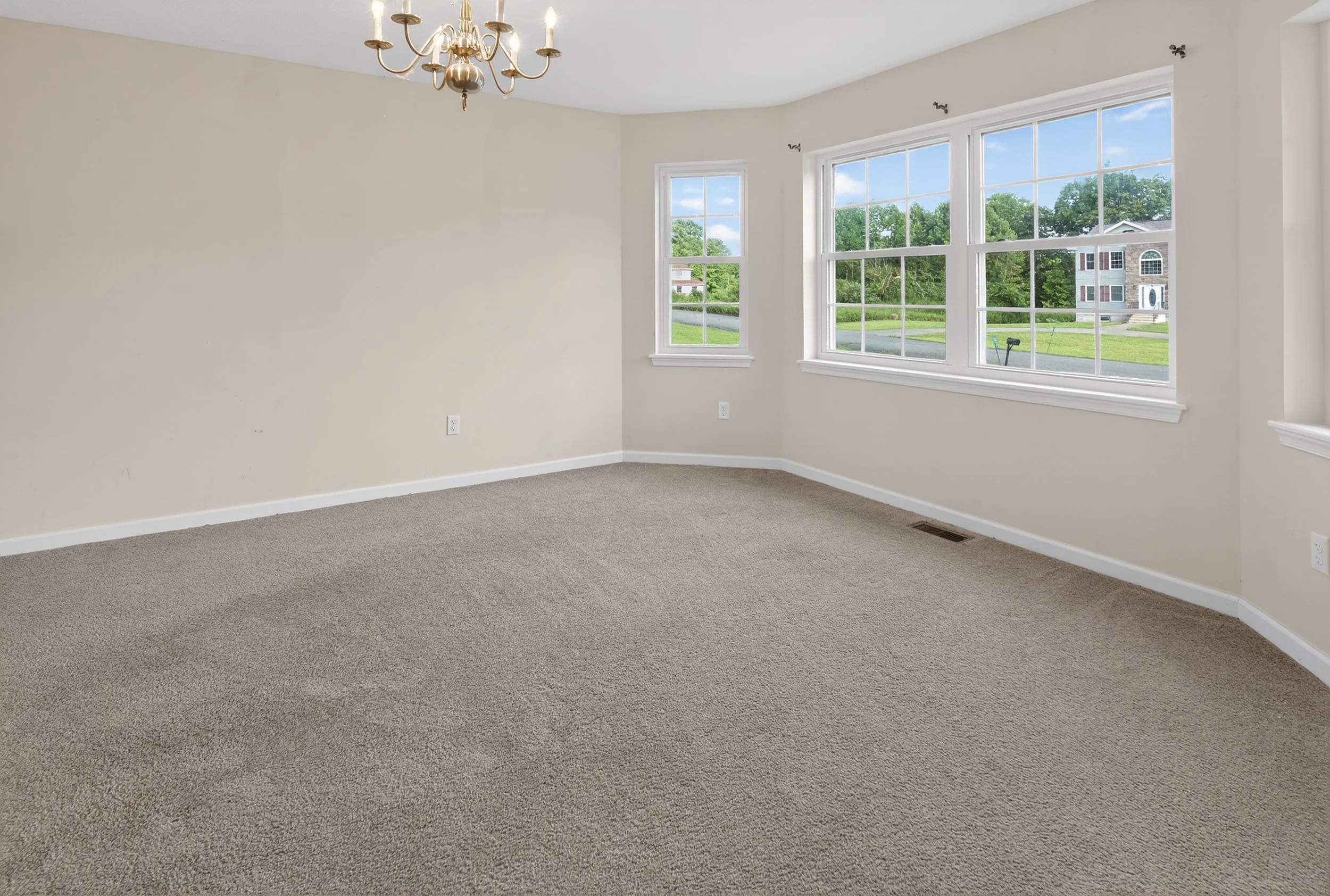 Empty living room with beige walls, gray carpet, large windows letting in natural light, and a brass chandelier hanging from the ceiling.