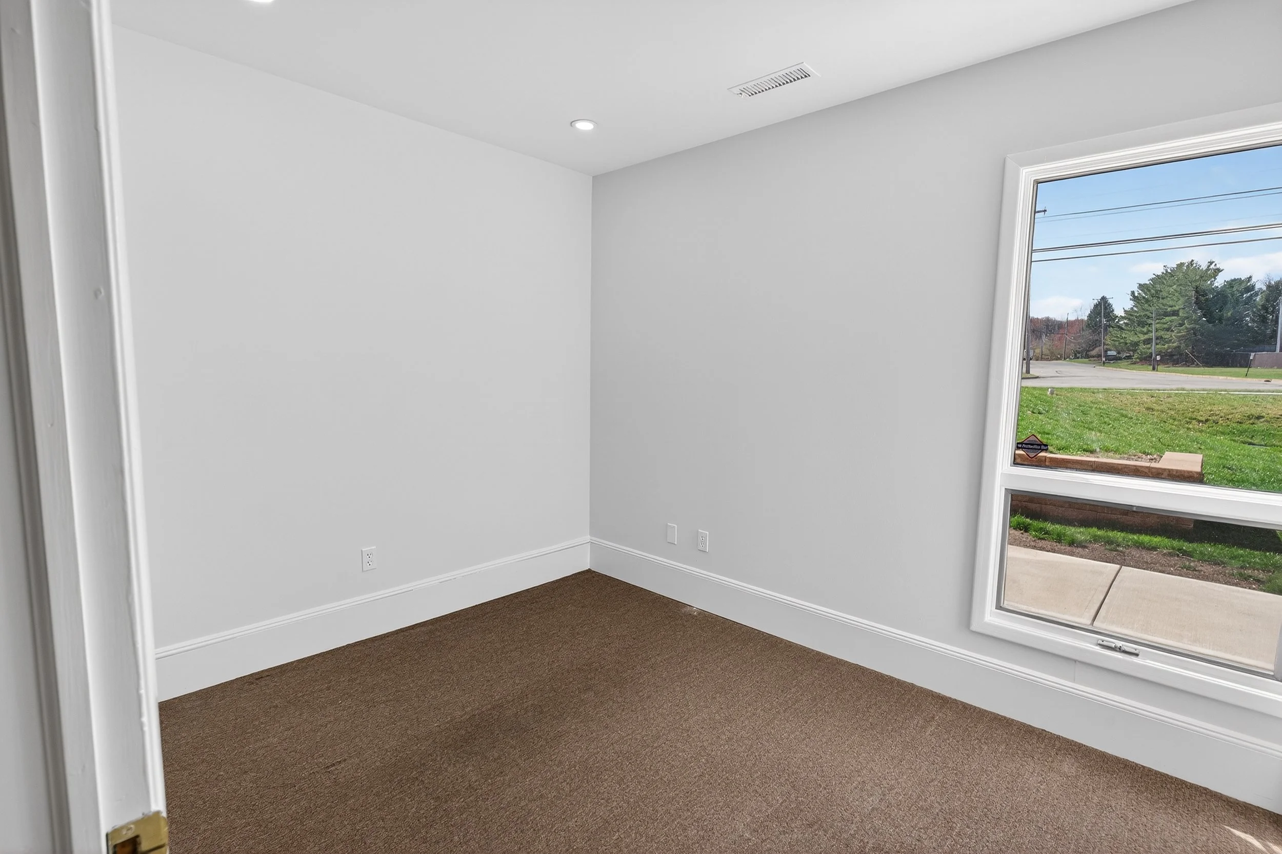 Empty room with white walls, brown carpet, large window showing a street and trees outside, and ceiling lights.