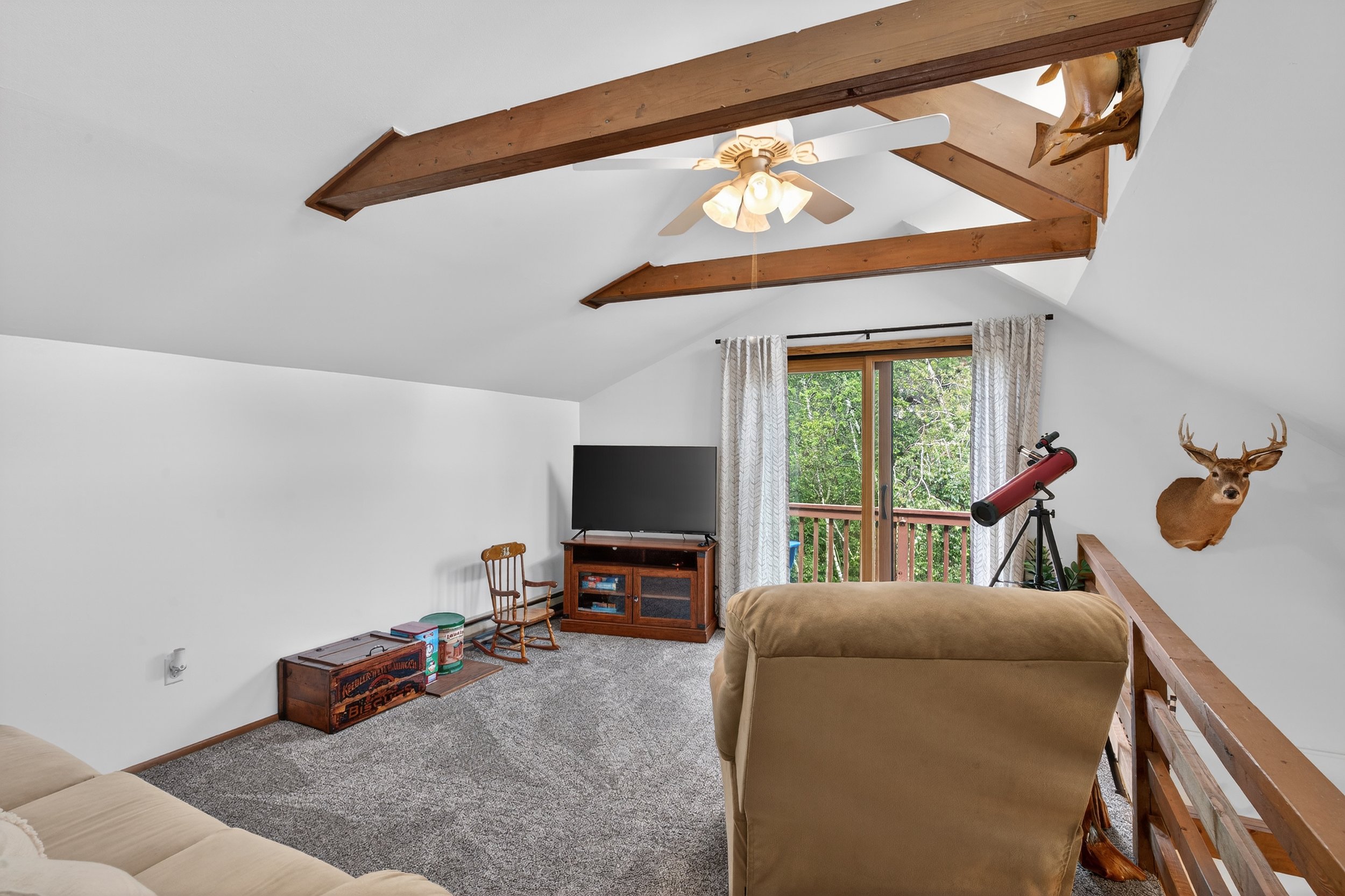 Living room with spindle chair, beige sofa, TV on a stand, telescope, mounted deer head, and wood beams on the ceiling, next to sliding glass door with curtains leading to a balcony overlooking trees.