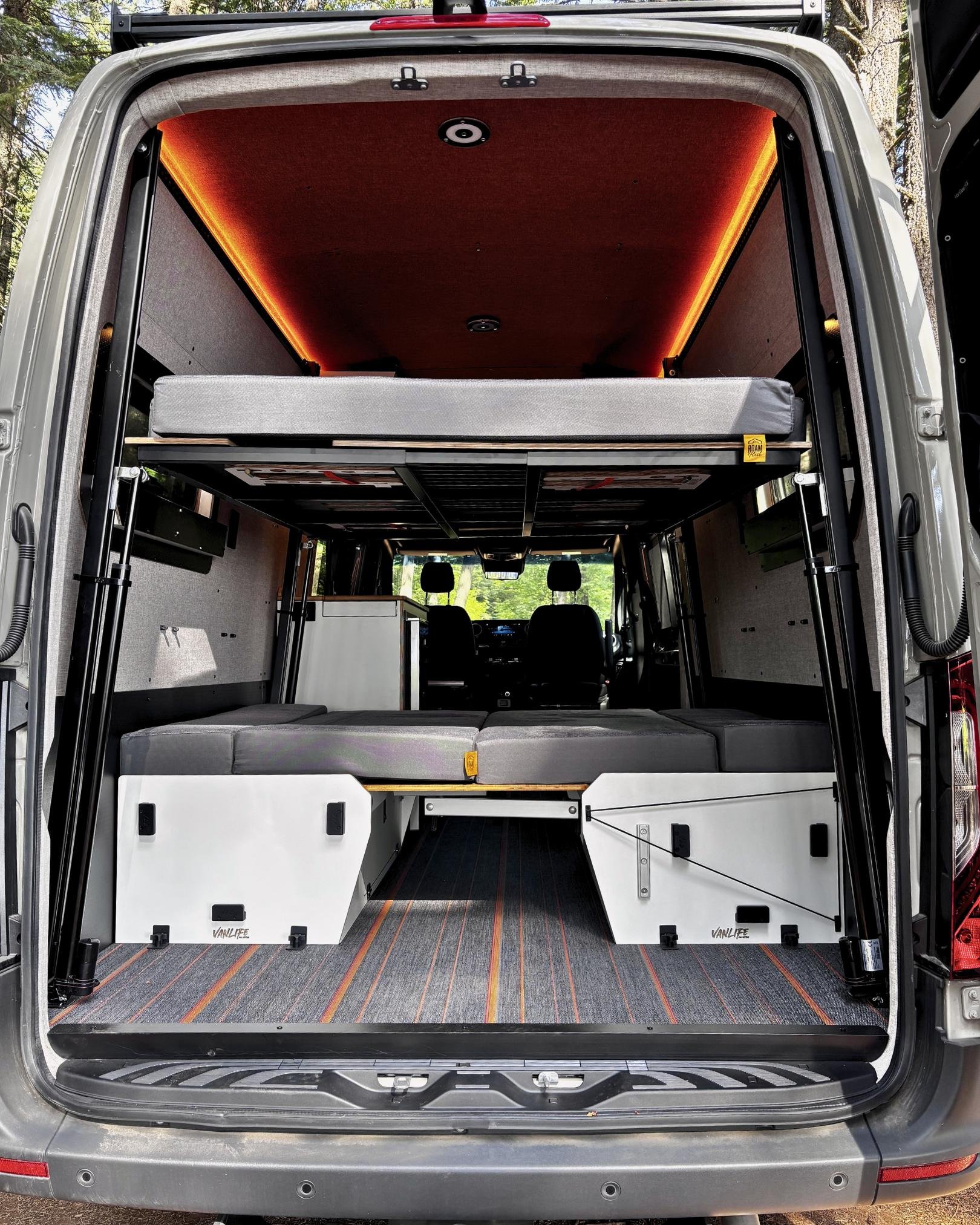 Extended Sprinter van conversion interior showing Elevation Station leveling bed raised and booth dinette with rear doors open to Oregon forest