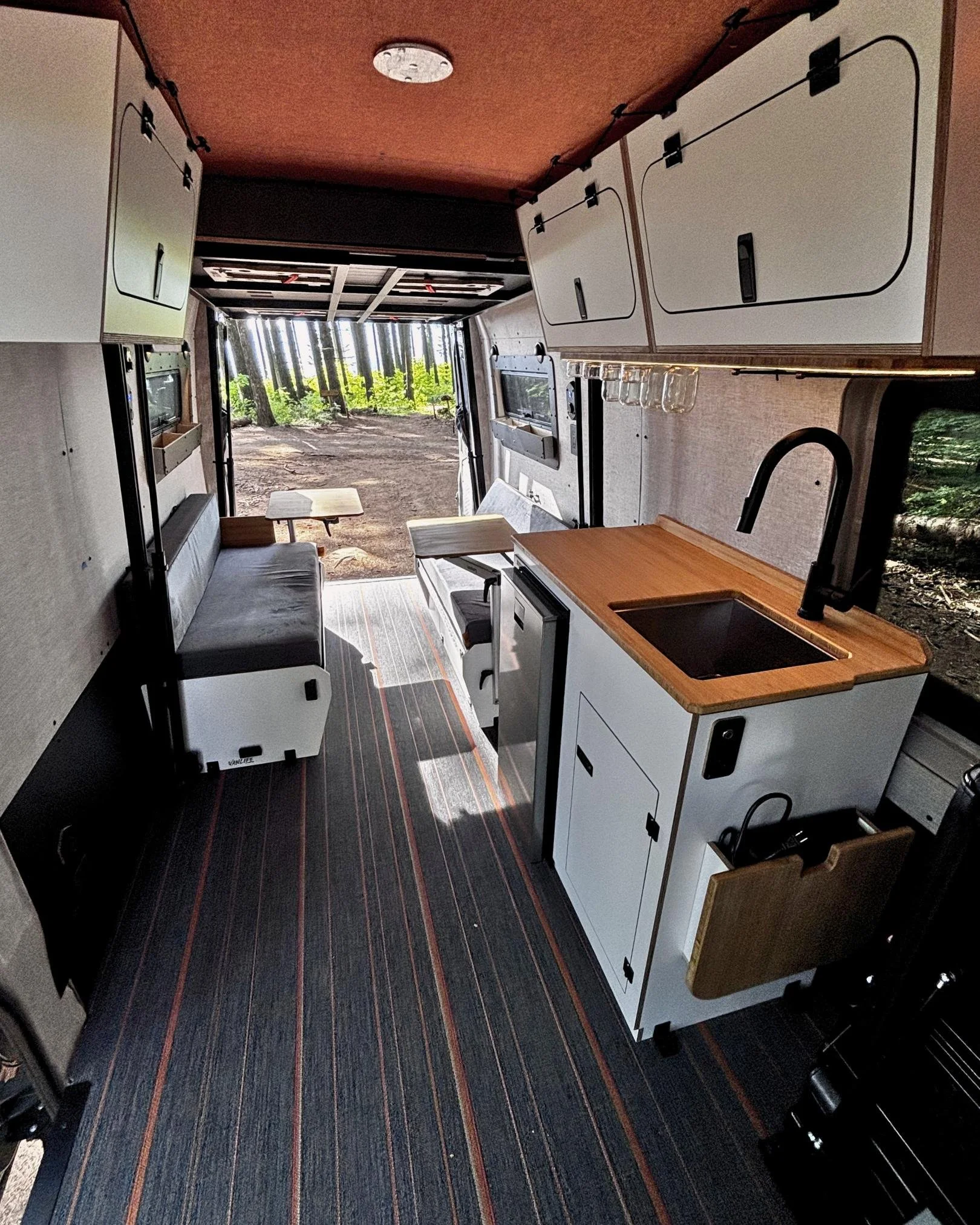 Interior of extended Sprinter van conversion showing galley kitchen, dinette seating and open rear doors to Oregon forest