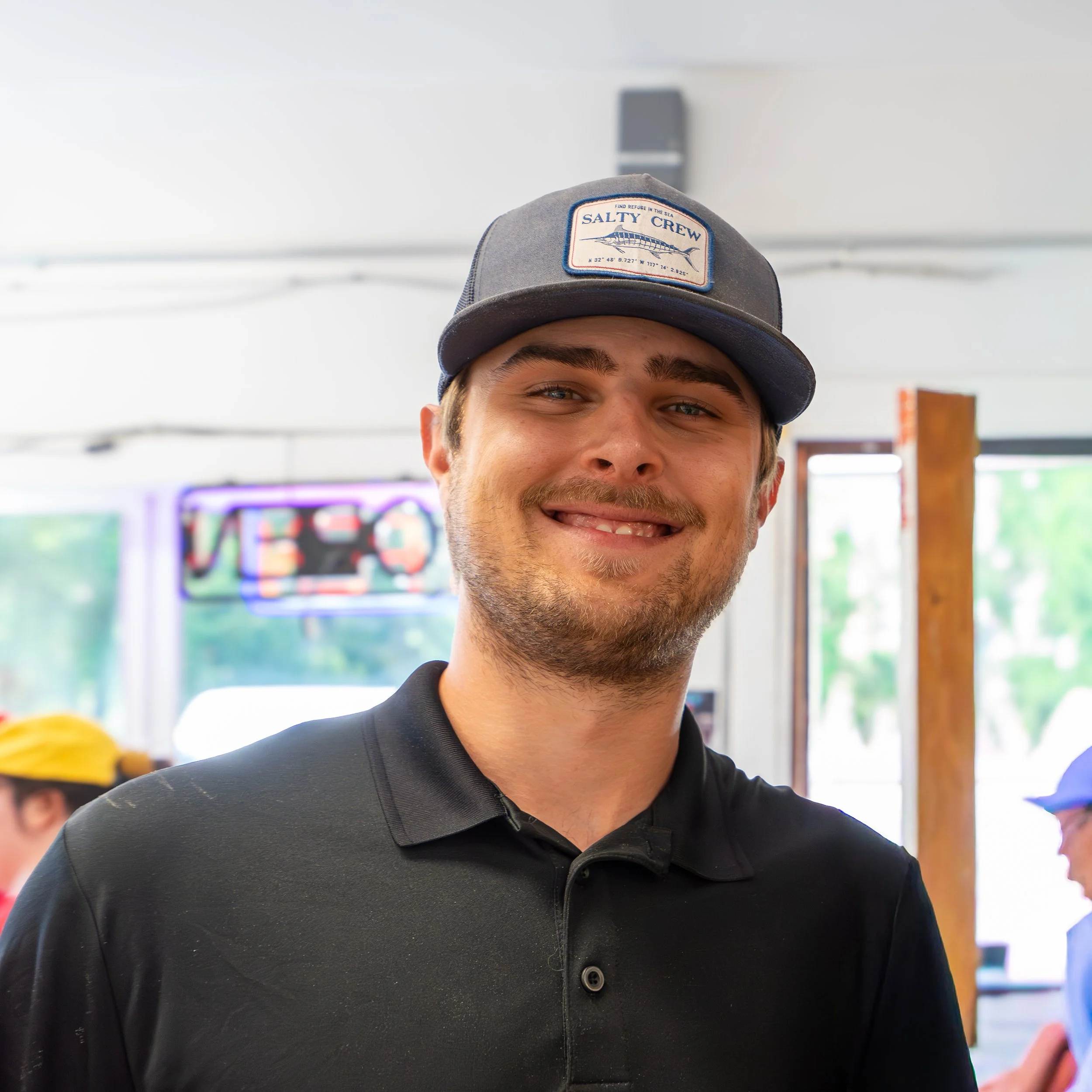 Close-up of a young man wearing a gray baseball cap with a "Salty Crew" patch, smiling, in an indoor setting with colorful background and other people.