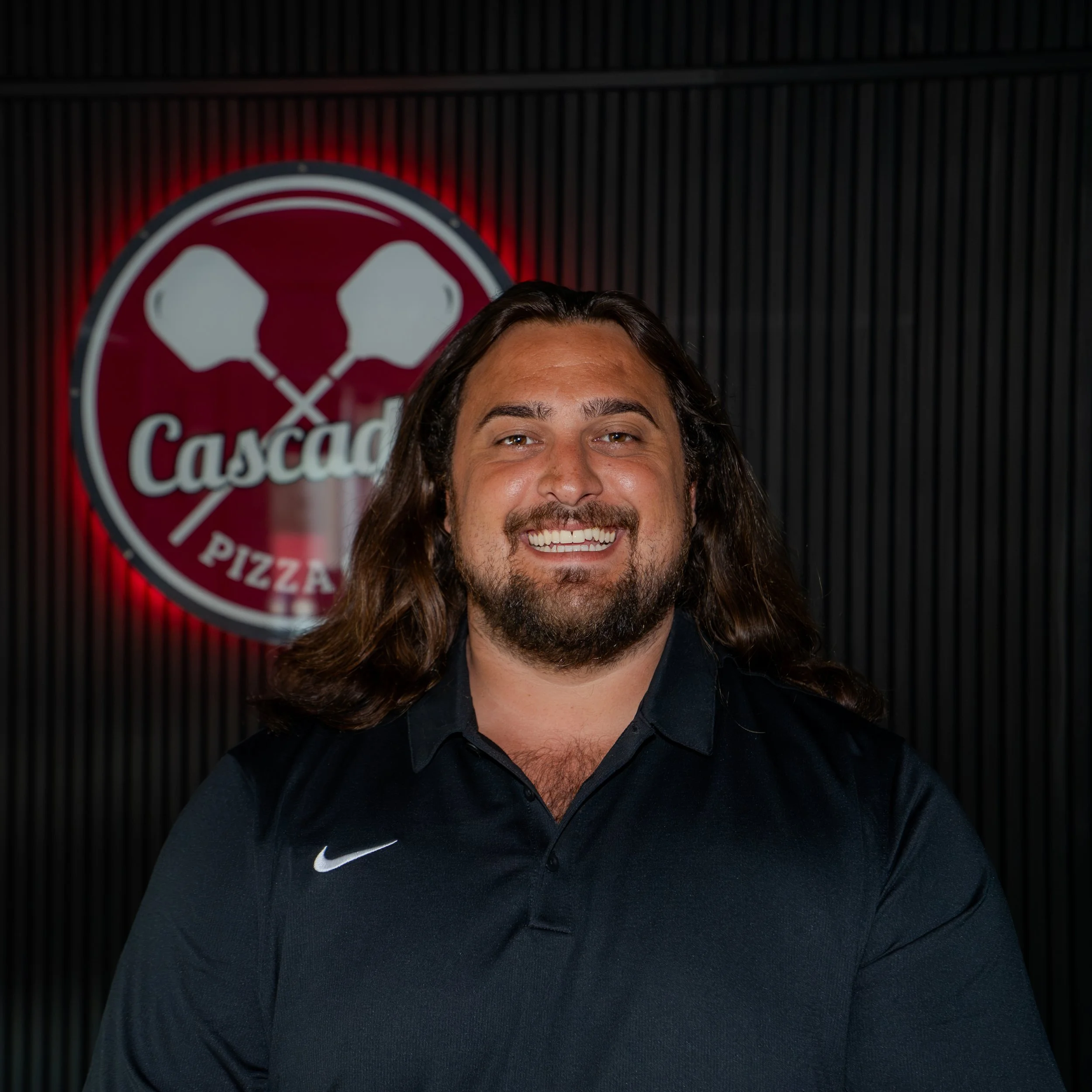 Close-up of a person with long brown hair smiling, wearing a black Nike jacket, with a red and white circular logo with crossed pizzerias in the background.