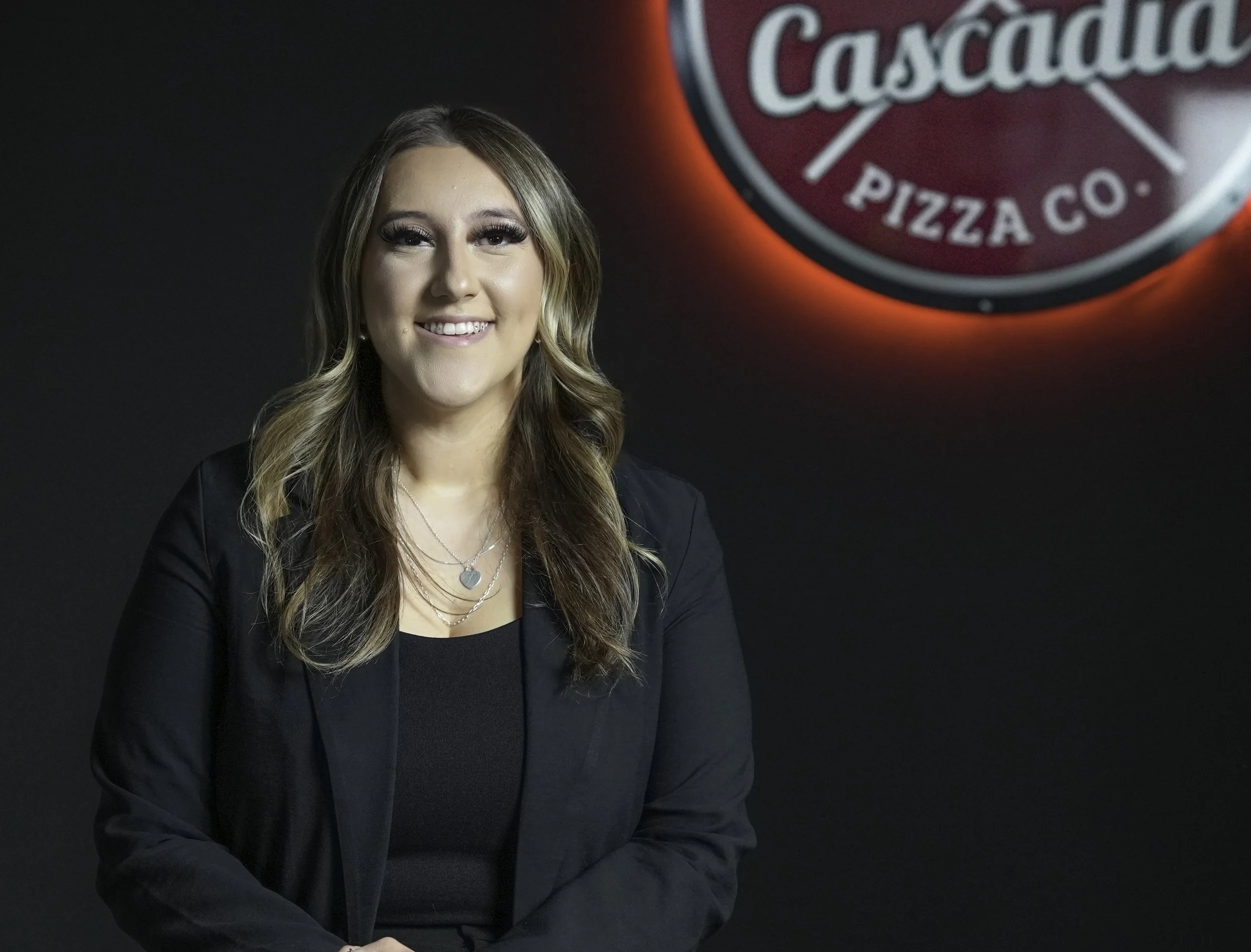 A young woman with long, wavy hair, wearing a black blazer and jewelry, smiling in front of a dark background with a large red illuminated sign reading 'Cascadia Pizza Co.'