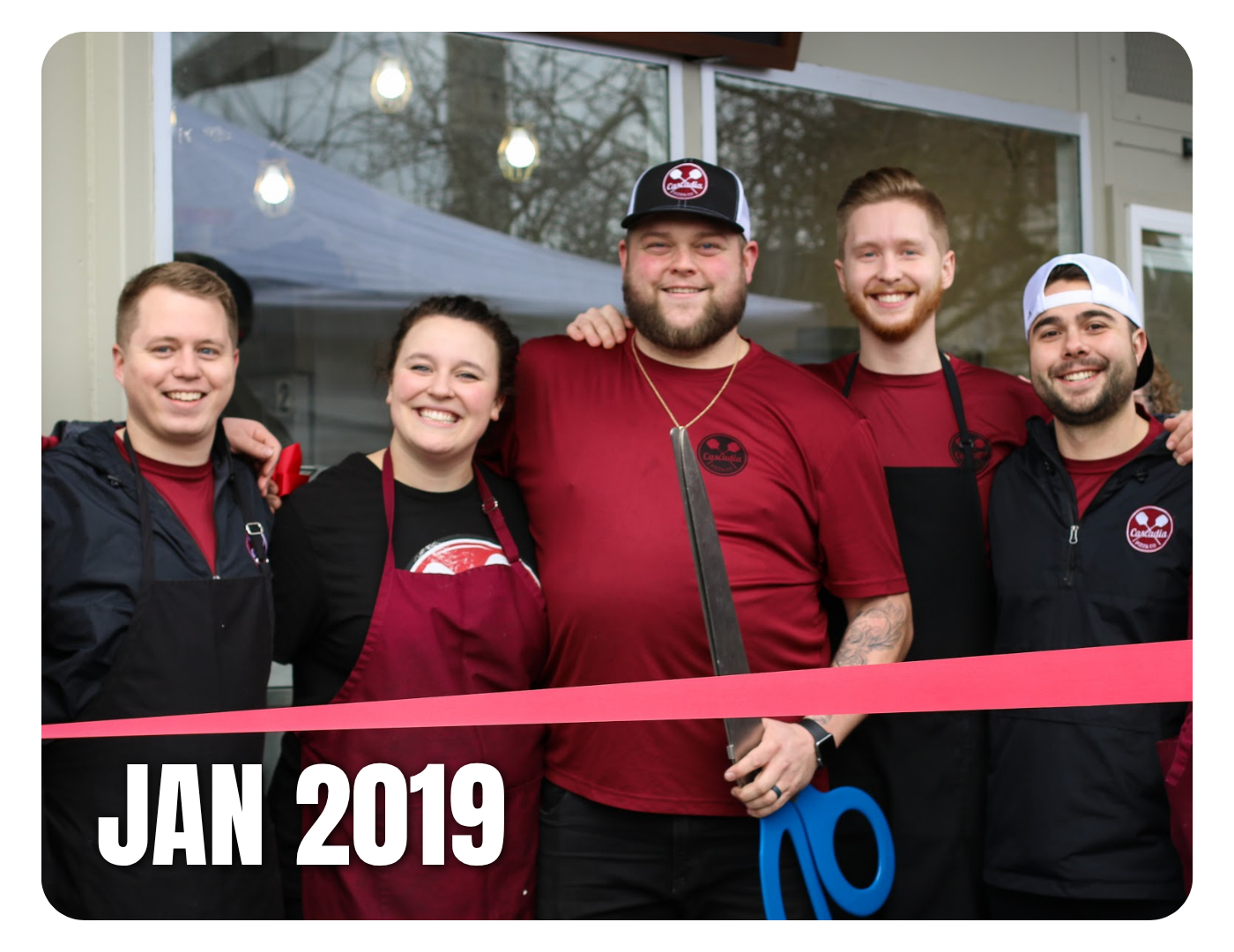 Group of five smiling people in aprons and team shirts standing behind a pink ribbon at a grand opening or event in January 2019, outside a building with a window and string lights