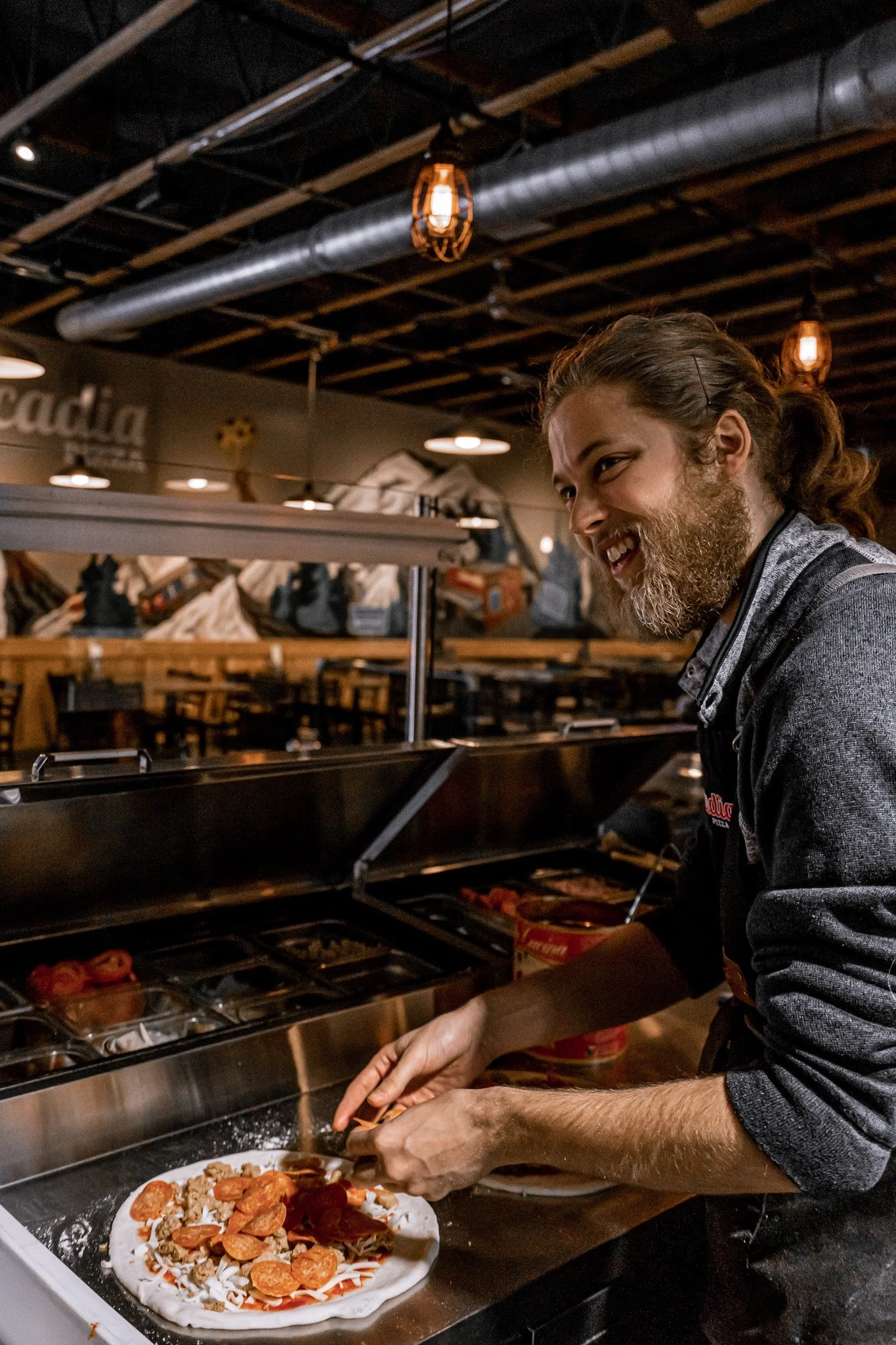 Man with long hair and beard preparing a pizza in a restaurant kitchen, with toppings including pepperoni and sausage. Wooden ceiling with exposed beams and hanging lights in the background.