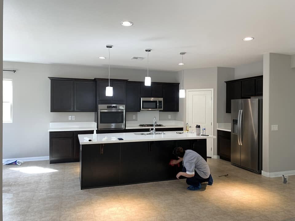 A person installing or cleaning cabinetry in a modern kitchen with stainless steel appliances, dark cabinets, a large kitchen island, and pendant lighting.