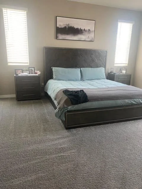 A bedroom with a large bed, dark wood nightstands on each side, gray headboard, light blue bedding, a black bag on the bed, and framed artwork above the bed. Two windows with blinds are on either side of the artwork.