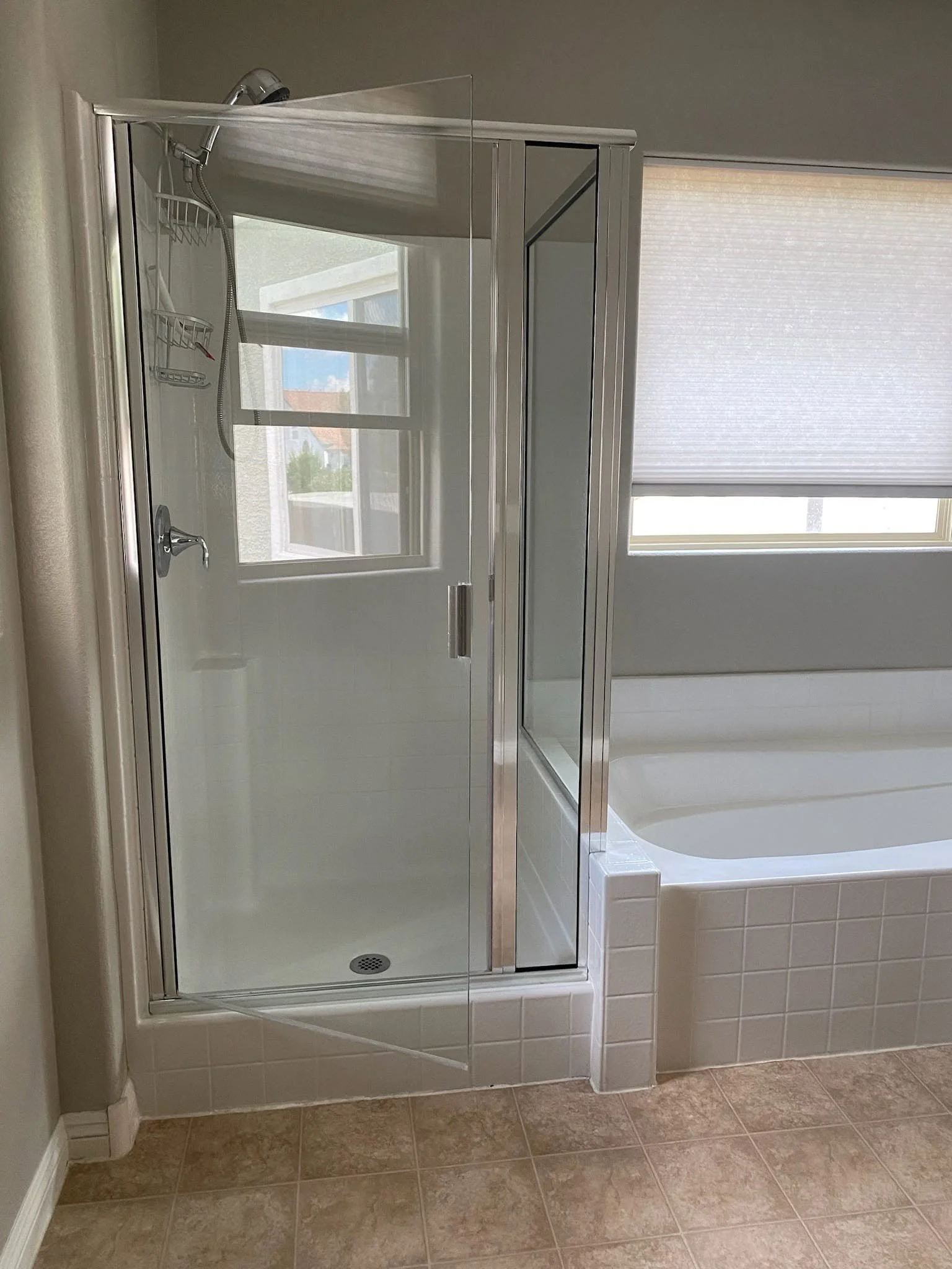 Bathroom shower area with glass door, handheld showerhead, and tiled walls, next to a soaking tub with a window in the background.