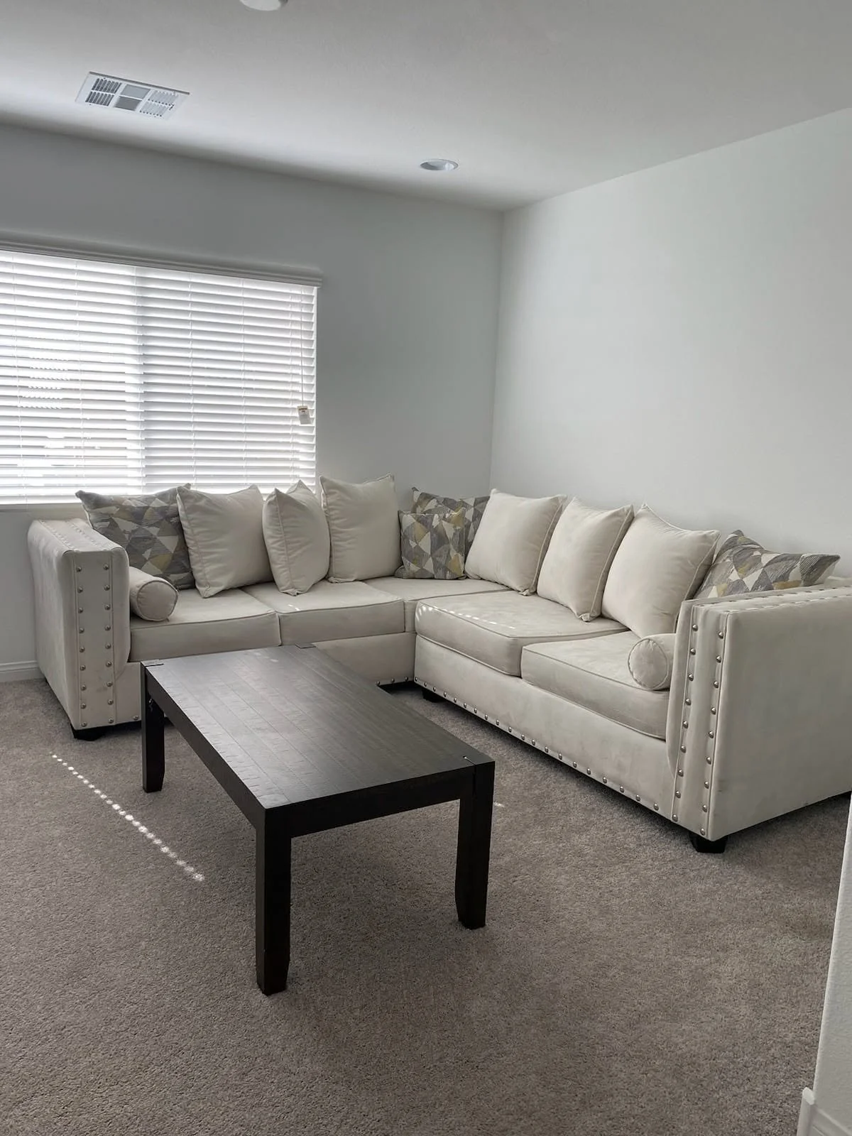 A living room with a beige sectional sofa with multiple pillows, a dark wooden coffee table, a window with blinds, and a beige carpet.