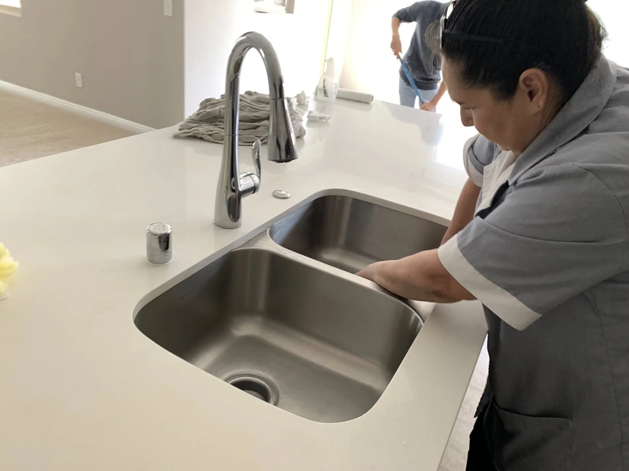 A person washing dishes at a kitchen sink, with another person in the background sweeping the floor.
