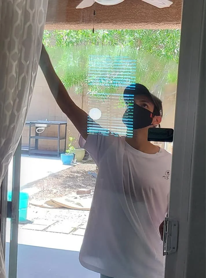 A boy wearing a face mask and a white T-shirt cleaning a glass door or window with a cloth or sponge, outdoors visible through the glass.