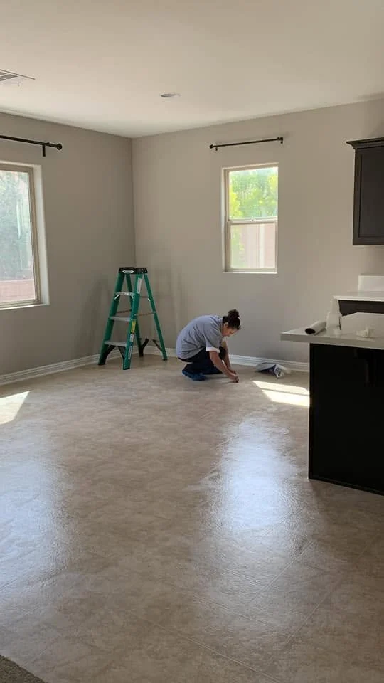 Person kneeling on the floor in a room, working on the flooring, with a step ladder nearby and windows providing natural light.