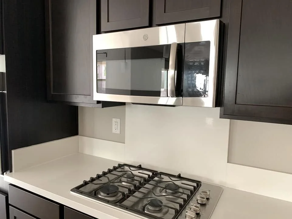 Stainless steel microwave oven installed above a four-burner gas stove in a modern kitchen with dark cabinets and a white countertop.