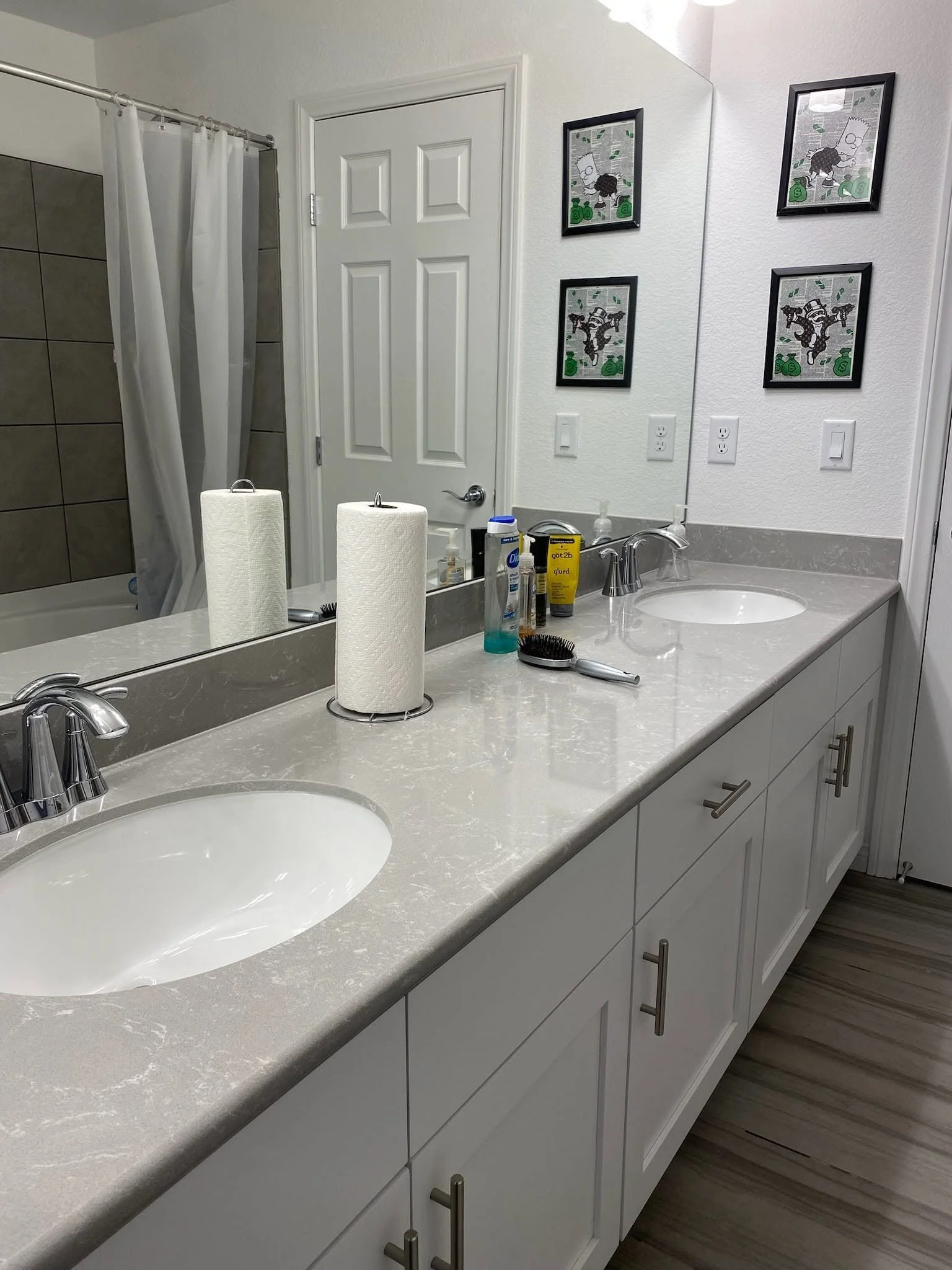 A bathroom vanity with a wide mirror, two sinks, and various toiletries including a paper towel roll, brushes, and bottles. The wall displays four framed pictures of cartoon characters.