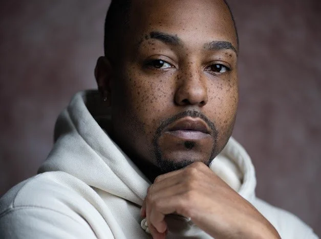 Close-up portrait of a man with dark skin, freckles, short hair, and a trimmed beard, wearing a white hoodie, against a dark blurred background.