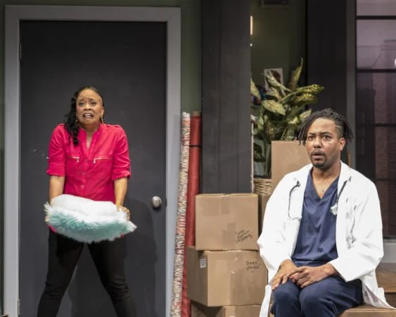 Two actors on stage performing a scene; the woman is standing, holding a fluffy pillow and appears surprised, while the man is sitting, wearing a doctor's coat and looking concerned, with boxes and plants in the background.