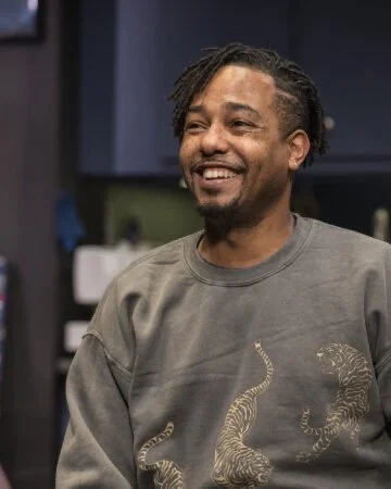 A smiling man with styled hair and a beard, wearing a gray sweatshirt with a tiger print, standing in a room with blue cabinets and a blurred background.