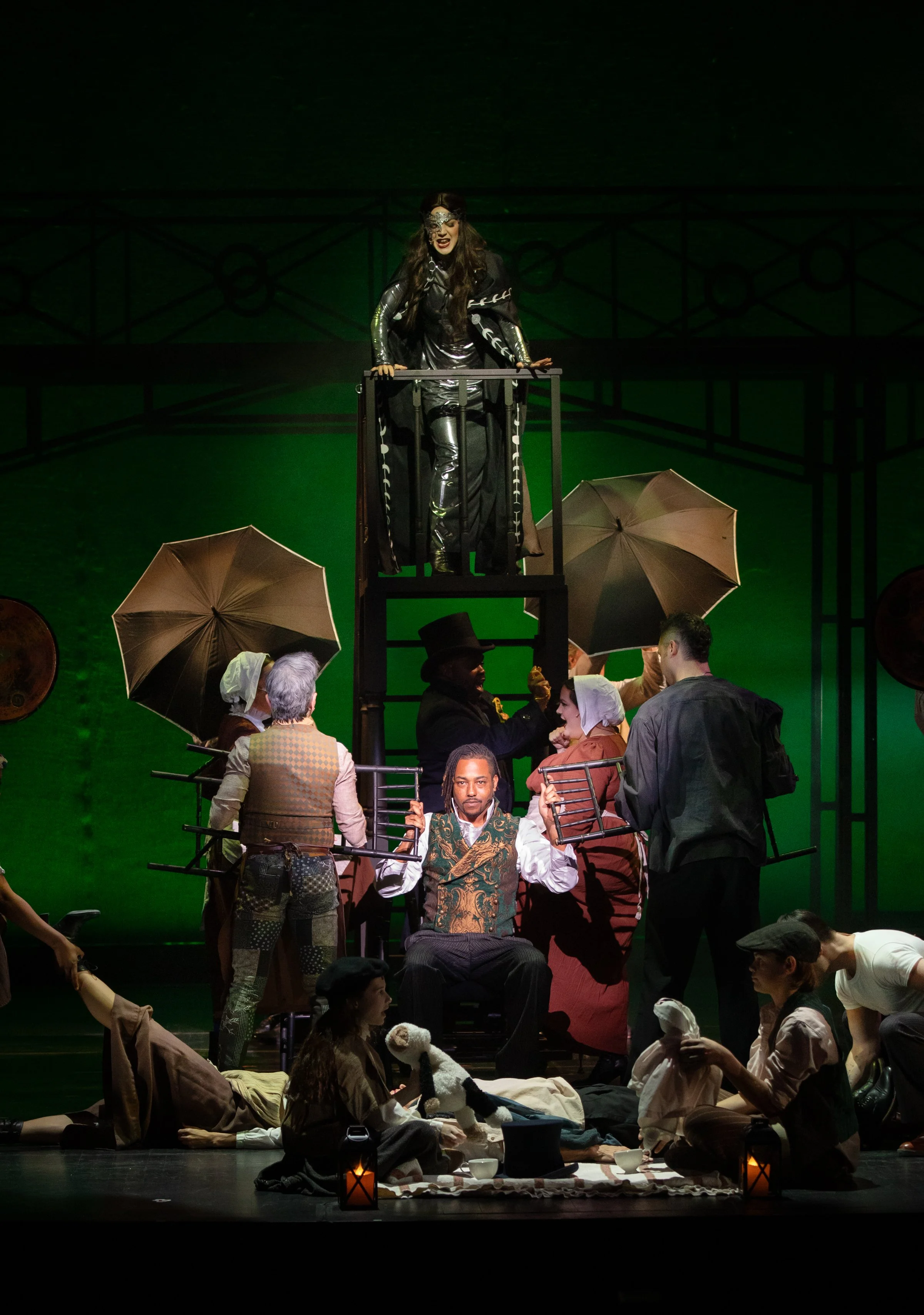 A theatrical scene with a woman dressed in black and metallic costume standing on a raised platform, surrounded by actors in period costumes holding umbrellas and props, with a green background.