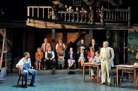 The scene depicts a theater stage with actors performing a play. There are people seated on stage and a man standing and speaking at a table. The backdrop resembles a two-story building, and some audience members are visible above the stage on a balcony.