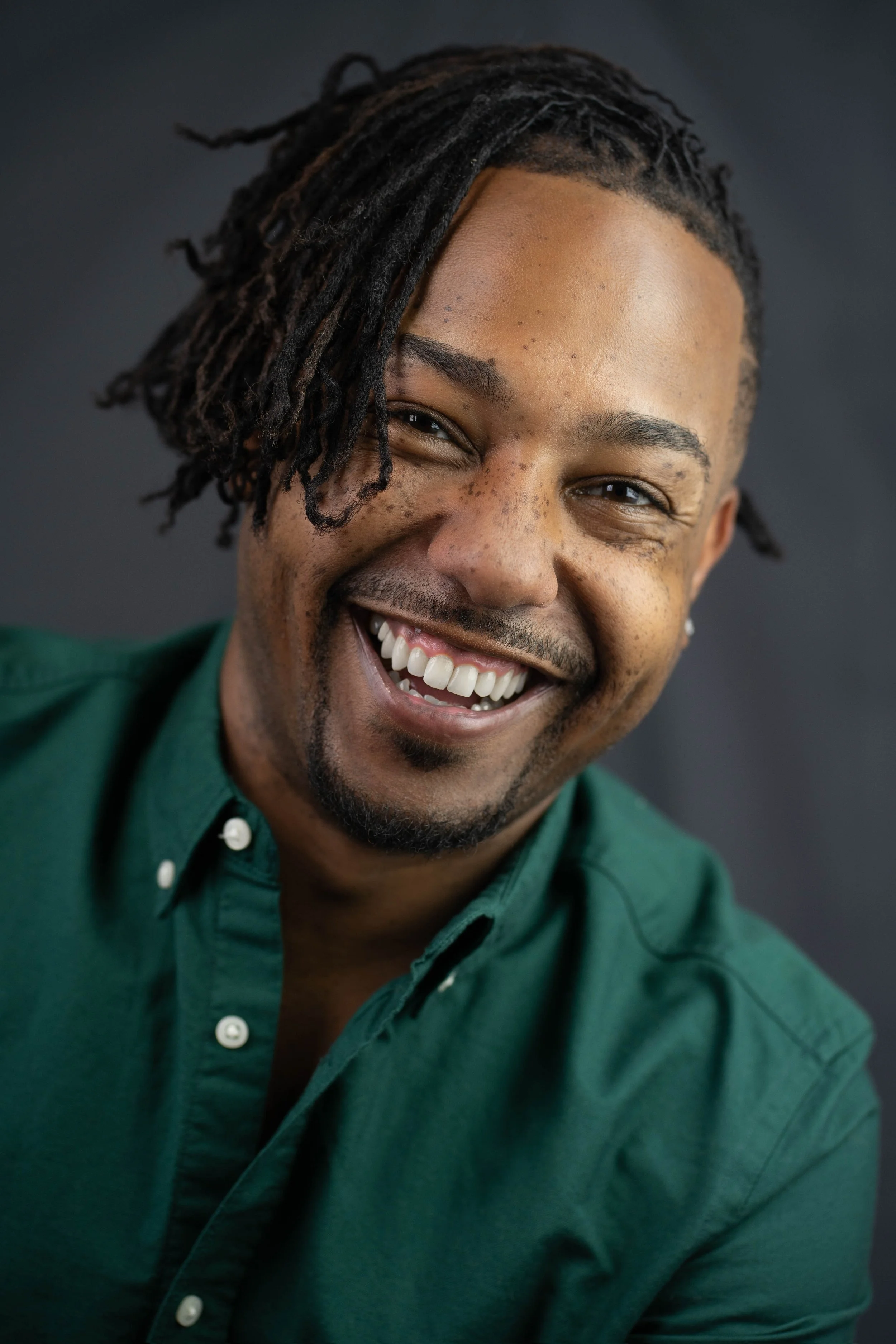 Close-up portrait of a smiling man with dreadlocks, wearing a green shirt, against a dark background.
