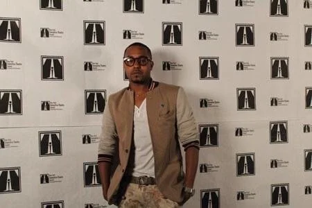 Man wearing glasses, beige and gray jacket, and white shirt standing in front of a wall with repeating graphic logos.