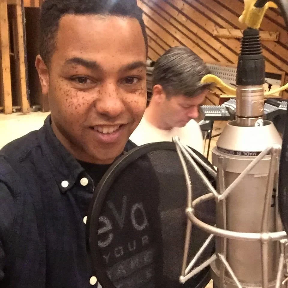 A young man with curly dark hair and freckles smiling at the camera in a recording studio, with another young man with straight hair focused on the music equipment in the background.