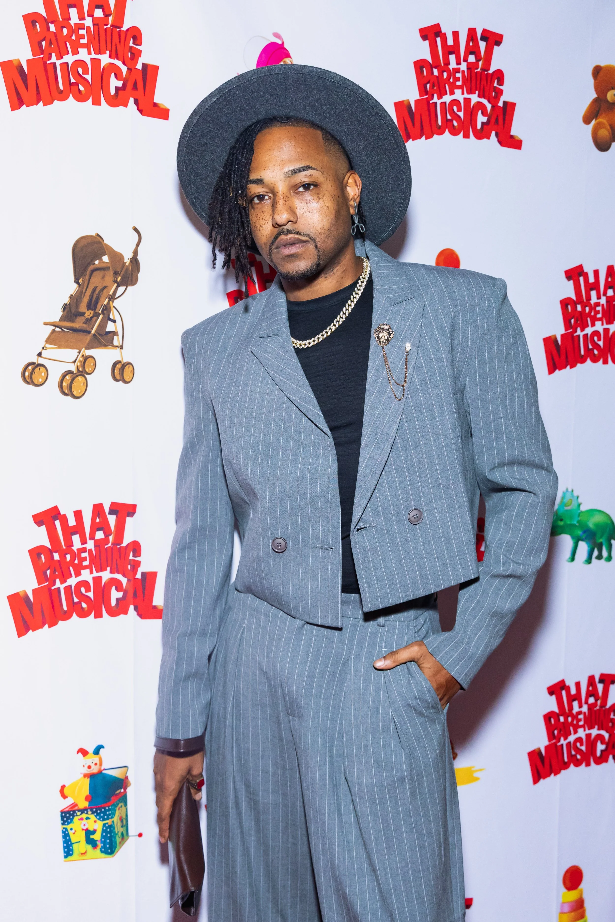 Person wearing a gray pinstripe suit, black shirt, silver necklace, earring, and a wide-brimmed gray hat, standing against a backdrop with colorful cartoon baby stroller and toy illustrations and red text reading "That Parenting Musical".