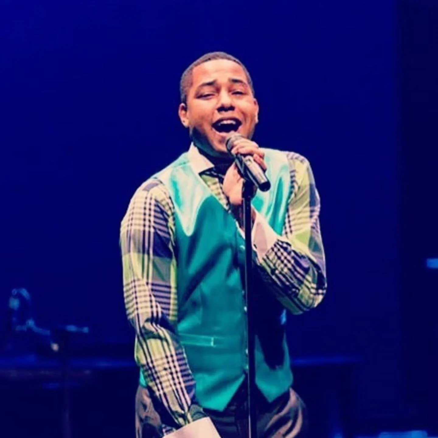 A young man singing into a microphone on stage, wearing a brightly colored patchwork vest and patterned shirt, with a dark background.