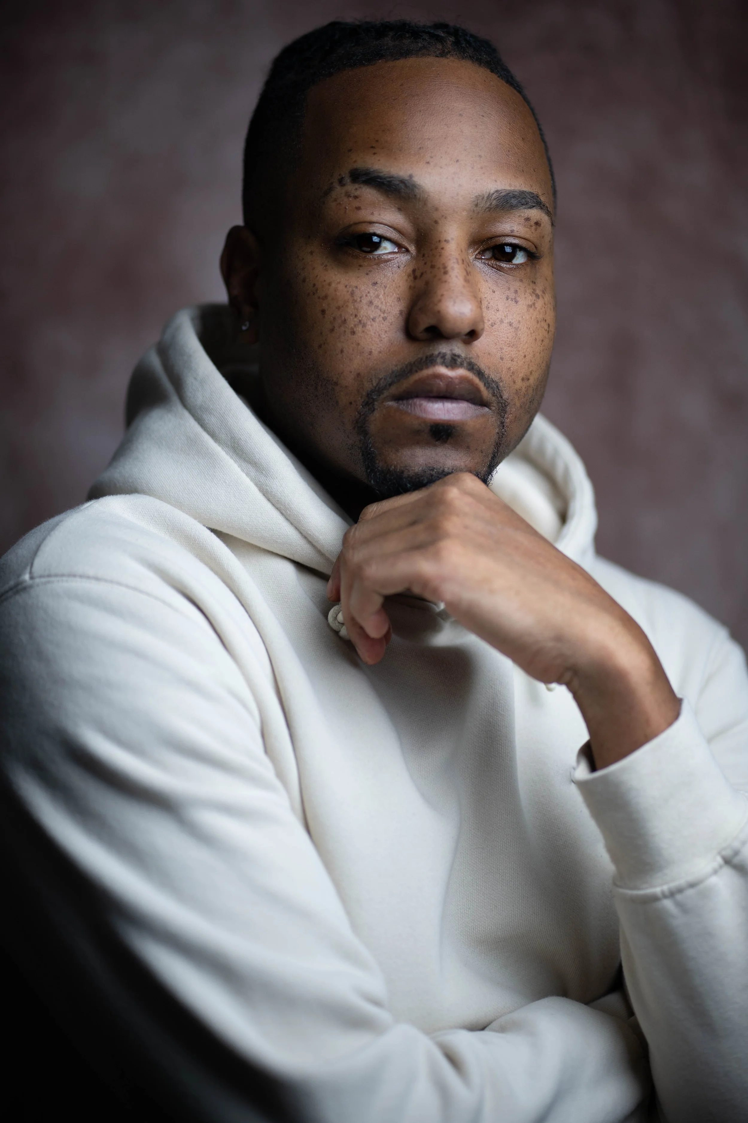Close-up of a young man with dark skin, freckles, and short dreadlocks, wearing a cream-colored hoodie, sitting with his hand near his chin, looking at the camera with a serious expression.