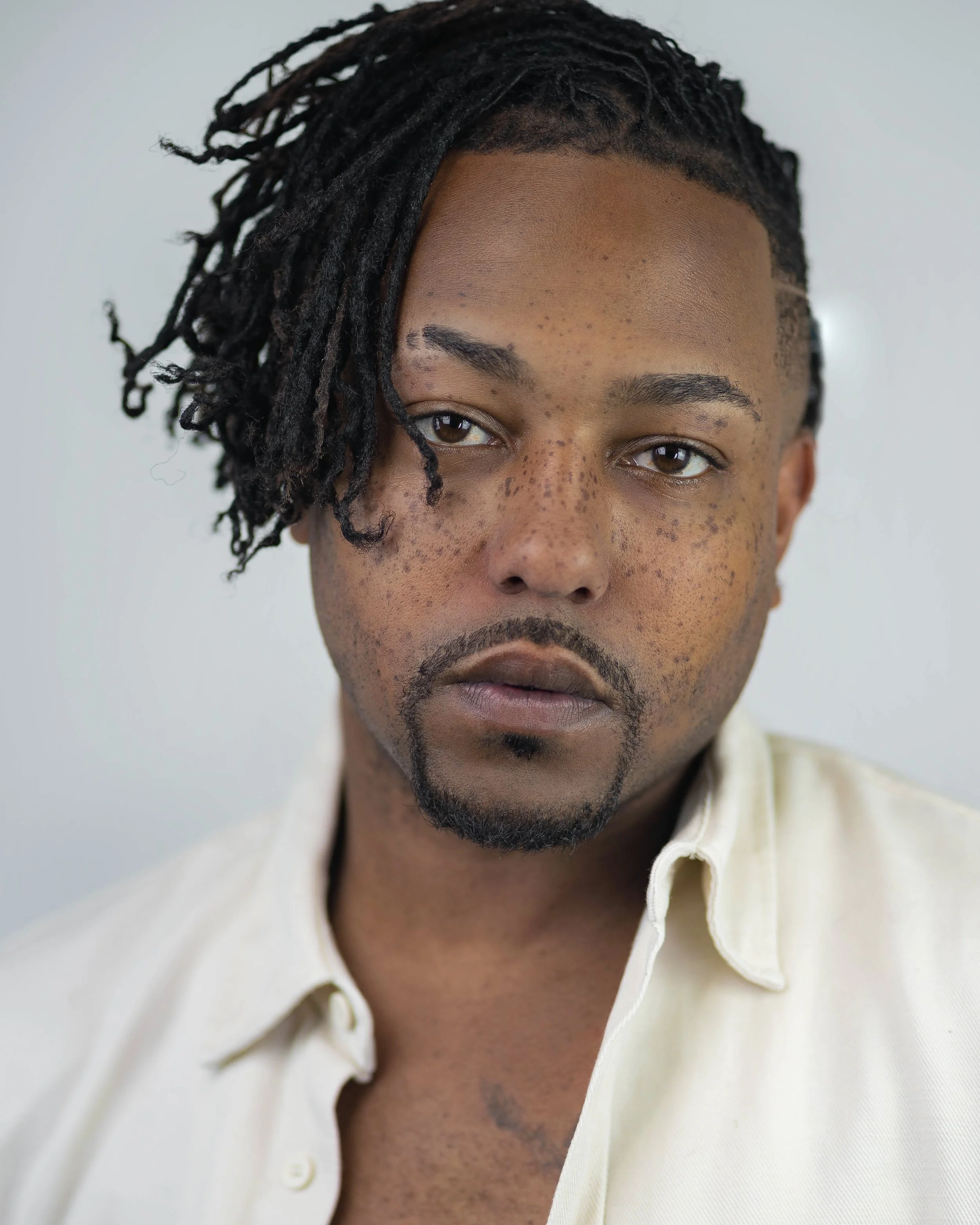 Close-up of a man with medium brown skin appearance, dark brown eyes, black facial hair, and short dreadlocks over his right eye, wearing a light-colored button-up shirt with the top button undone, against a plain light background.
