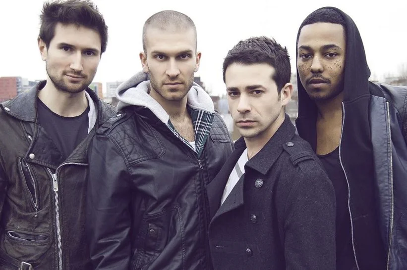 Four young men standing outdoors in a city, wearing dark jackets, with serious expressions.