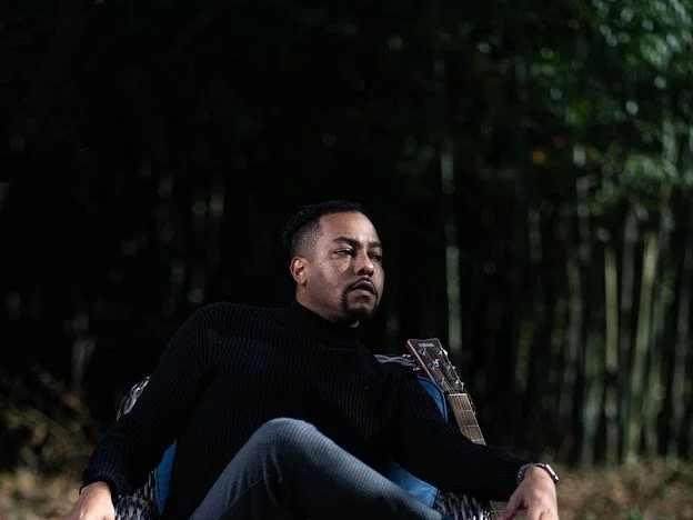 A man sitting outdoors in a dark, wooded area, wearing a black sweater, with a calm and contemplative expression.