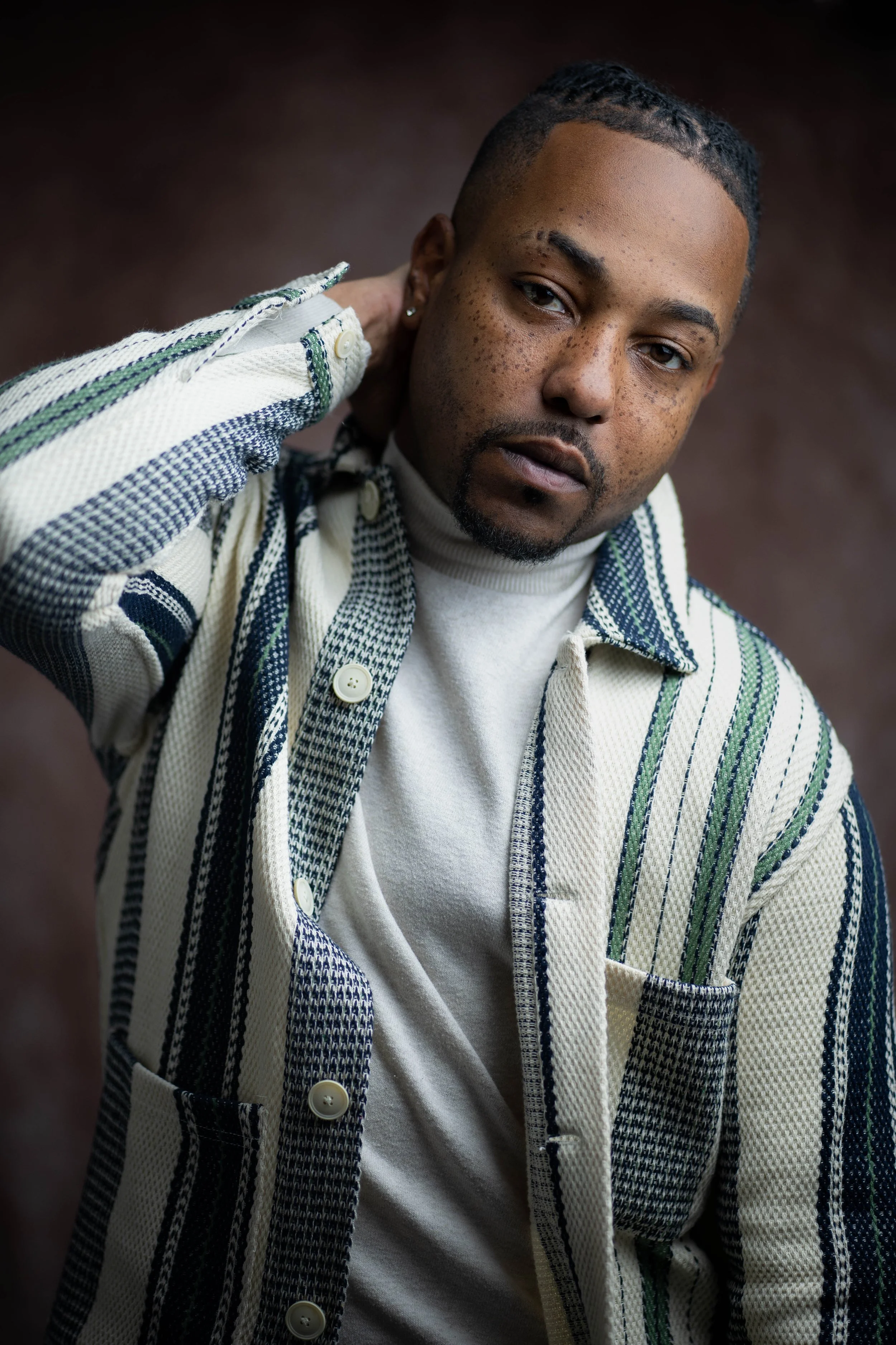 A young man with short hair, freckles, and a trimmed beard is posing with one hand behind his neck, wearing a patterned striped button-up shirt over a light-colored turtleneck, against a dark background.