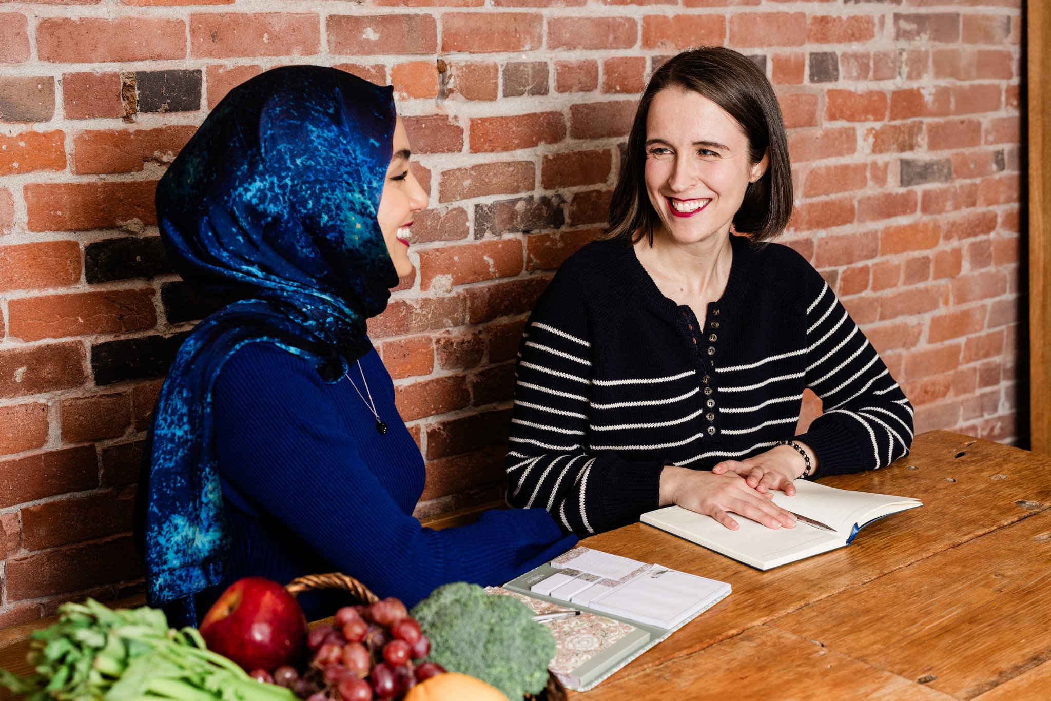 Two women sitting at a wooden table, engaging in conversation, with a brick wall in the background. The woman on the left is wearing a blue patterned headscarf and a blue top, with a basket of fruits and vegetables on the table. The woman on the right has short brown hair, is smiling, and has an open notebook in front of her.