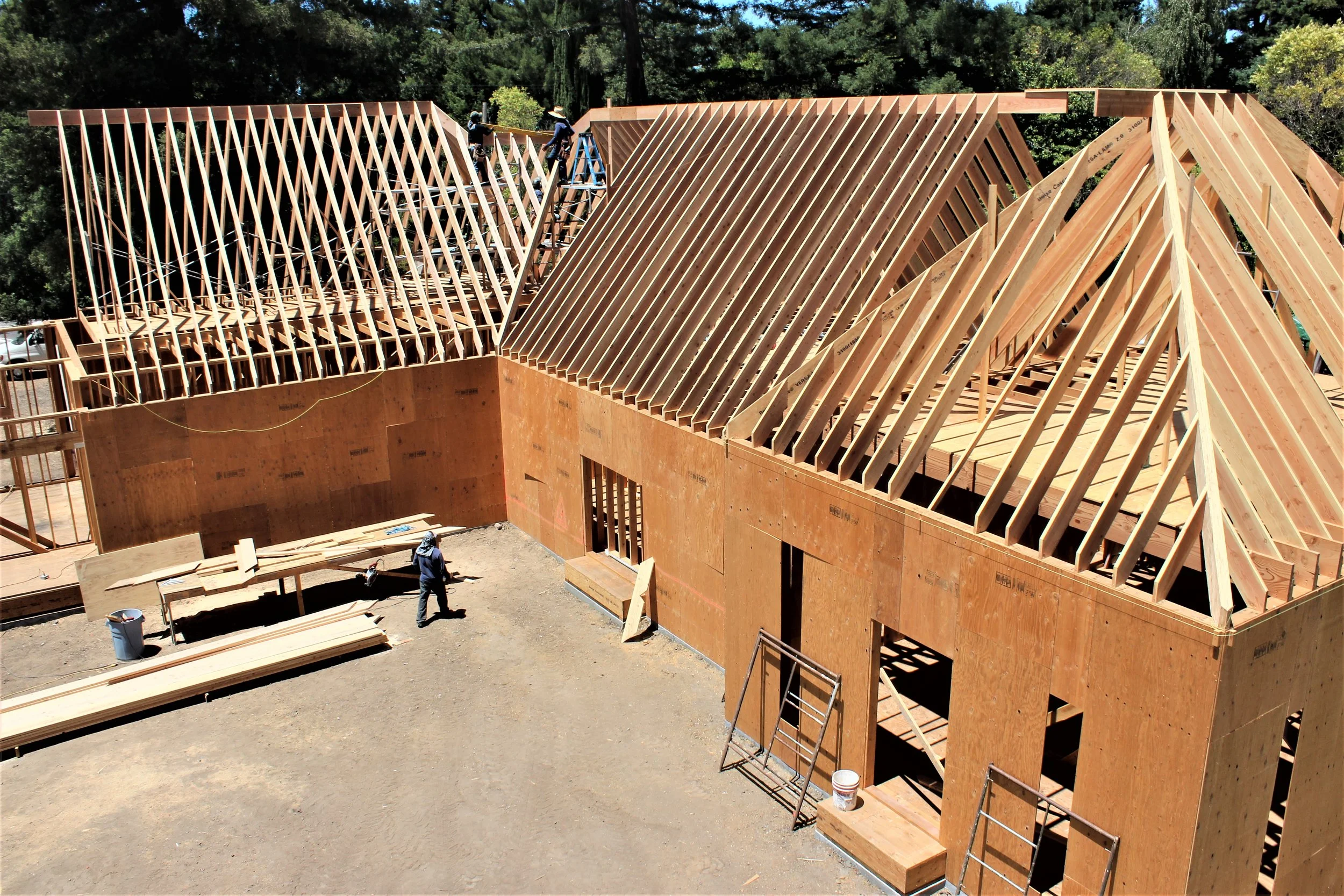 Construction site of a house with wooden framing and roof trusses, workers on scaffolding, and building materials on the ground.