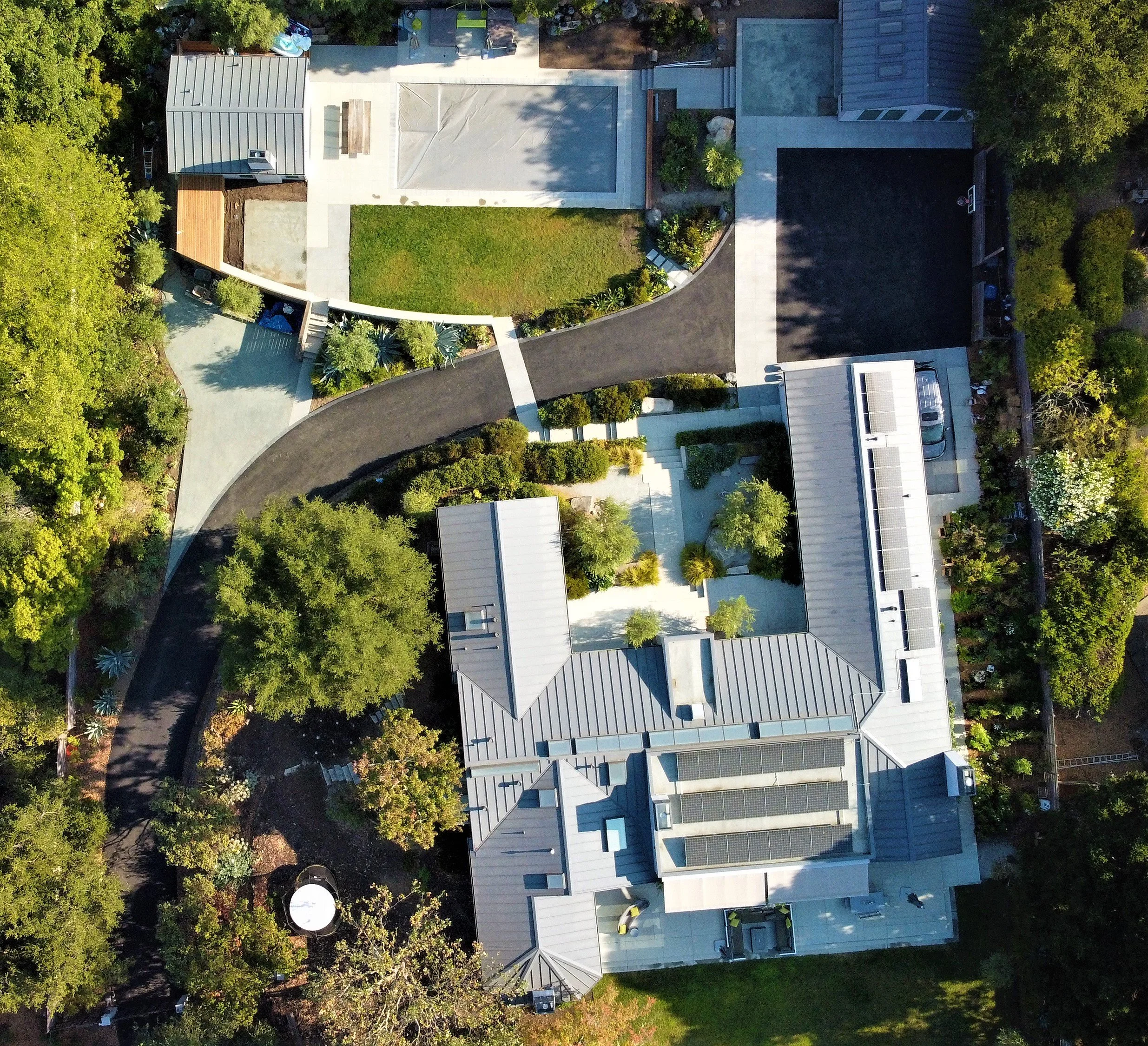 Aerial view of a modern house with a metal roof, surrounded by trees, a driveway, and landscaped garden areas, including a basketball court and paved walkways.
