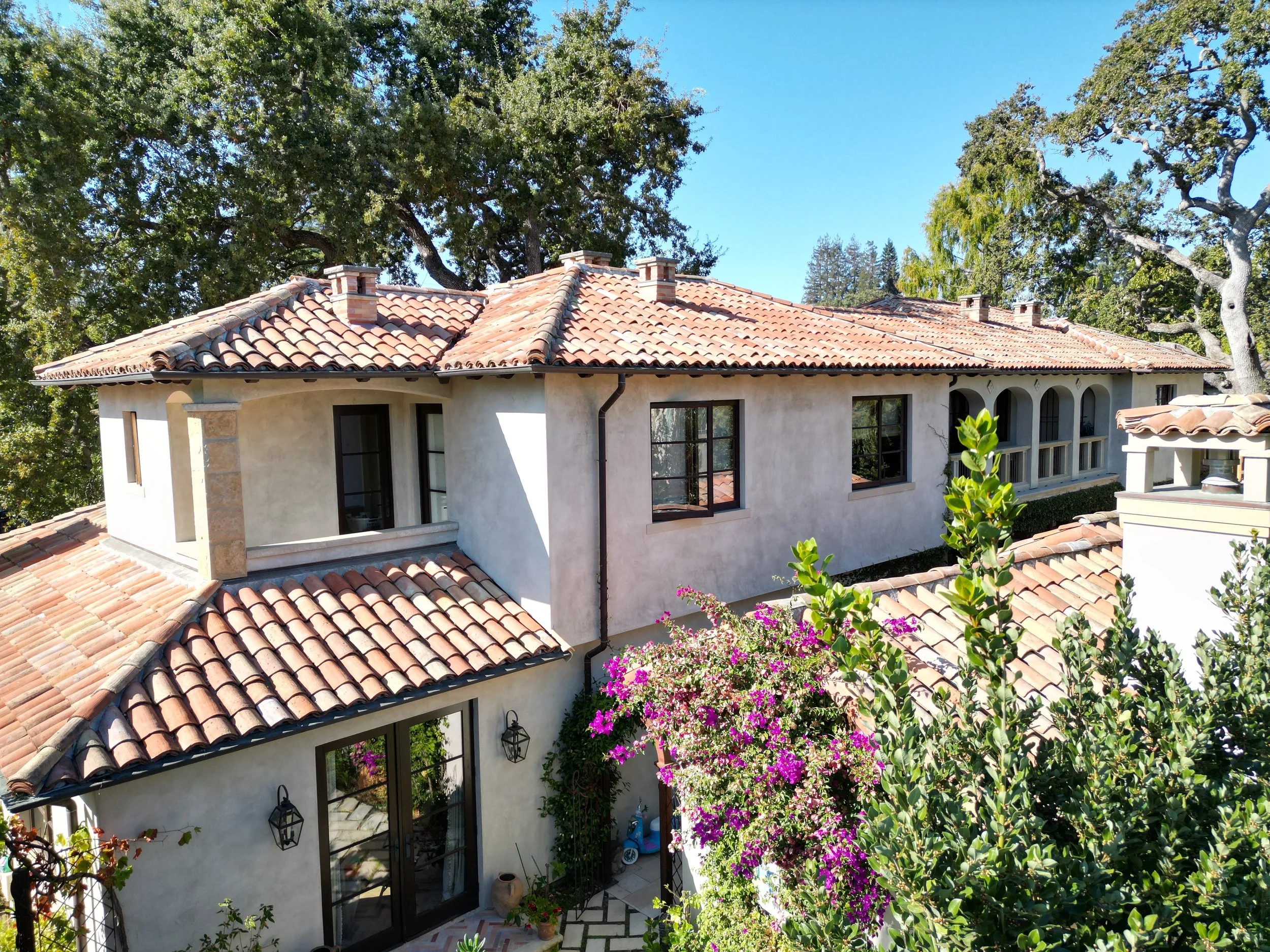 A Mediterranean-style house with a red tile roof, white stucco walls, and multiple black-framed windows, surrounded by green trees and flowering bushes.