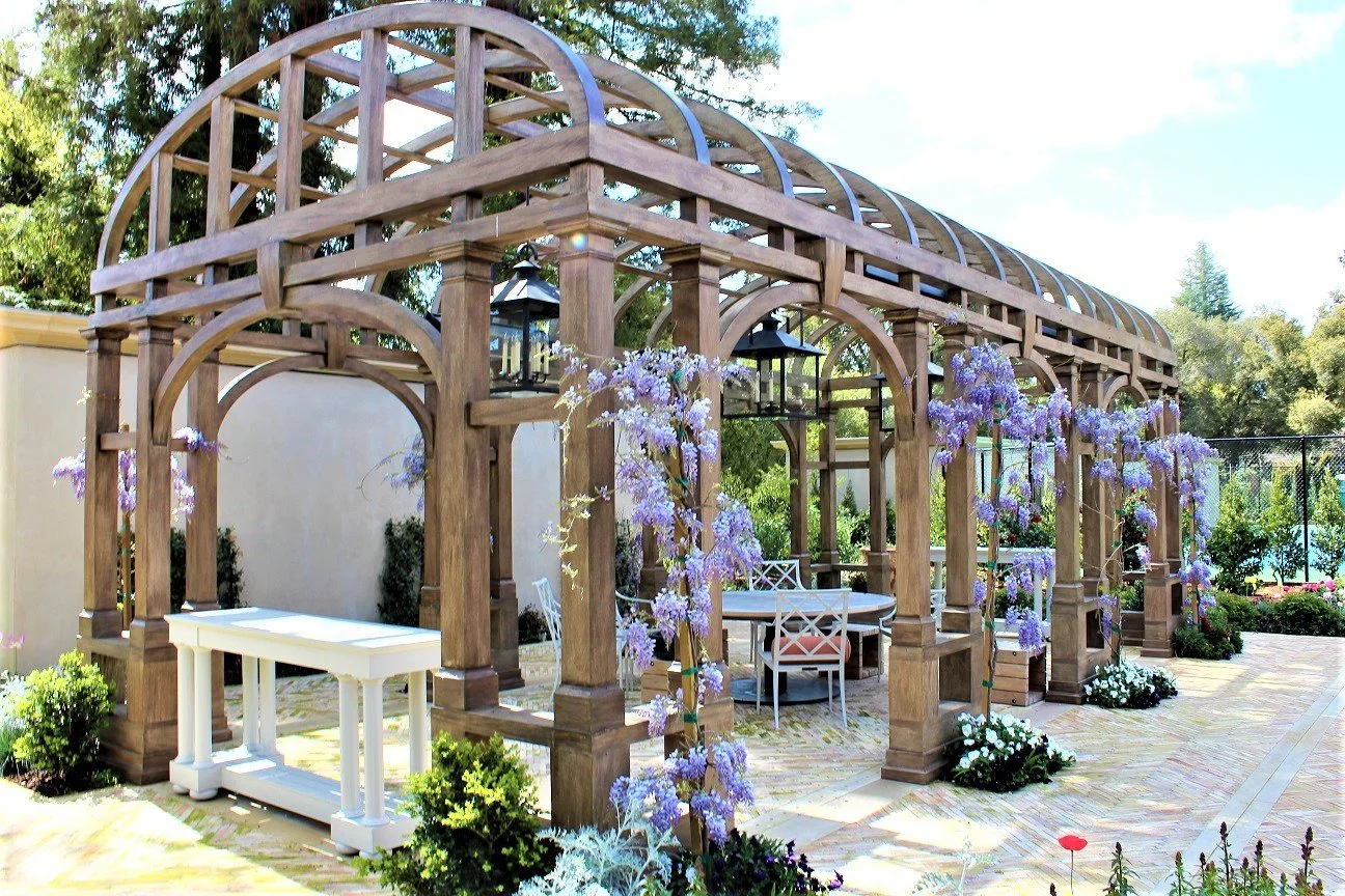 A wooden pergola with purple flowering vines, outdoor furniture, and hanging lanterns in a garden setting.