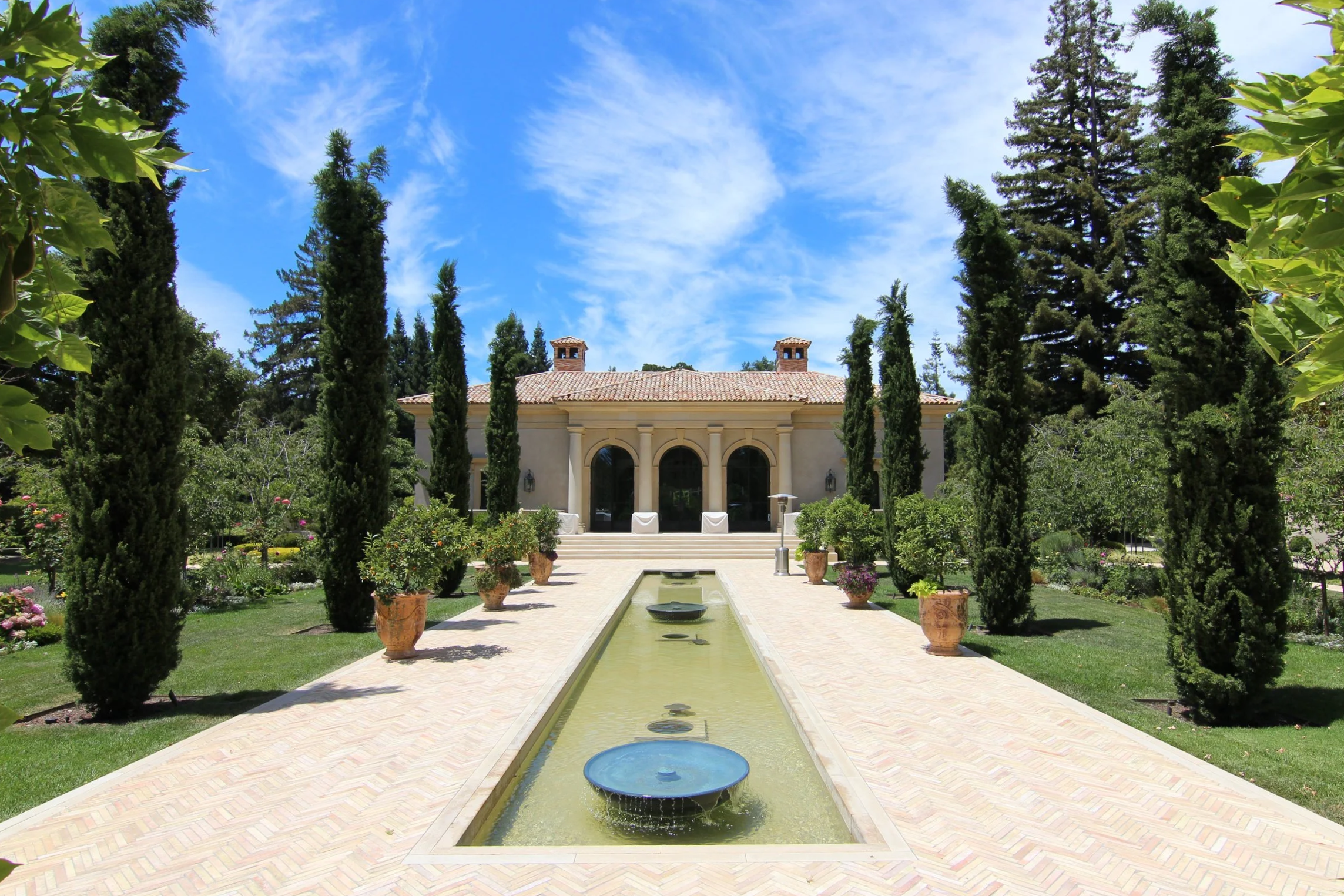 Front view of a luxurious house with a garden, walkways, a long water feature, tall cypress trees, potted plants, and blue sky with some clouds.
