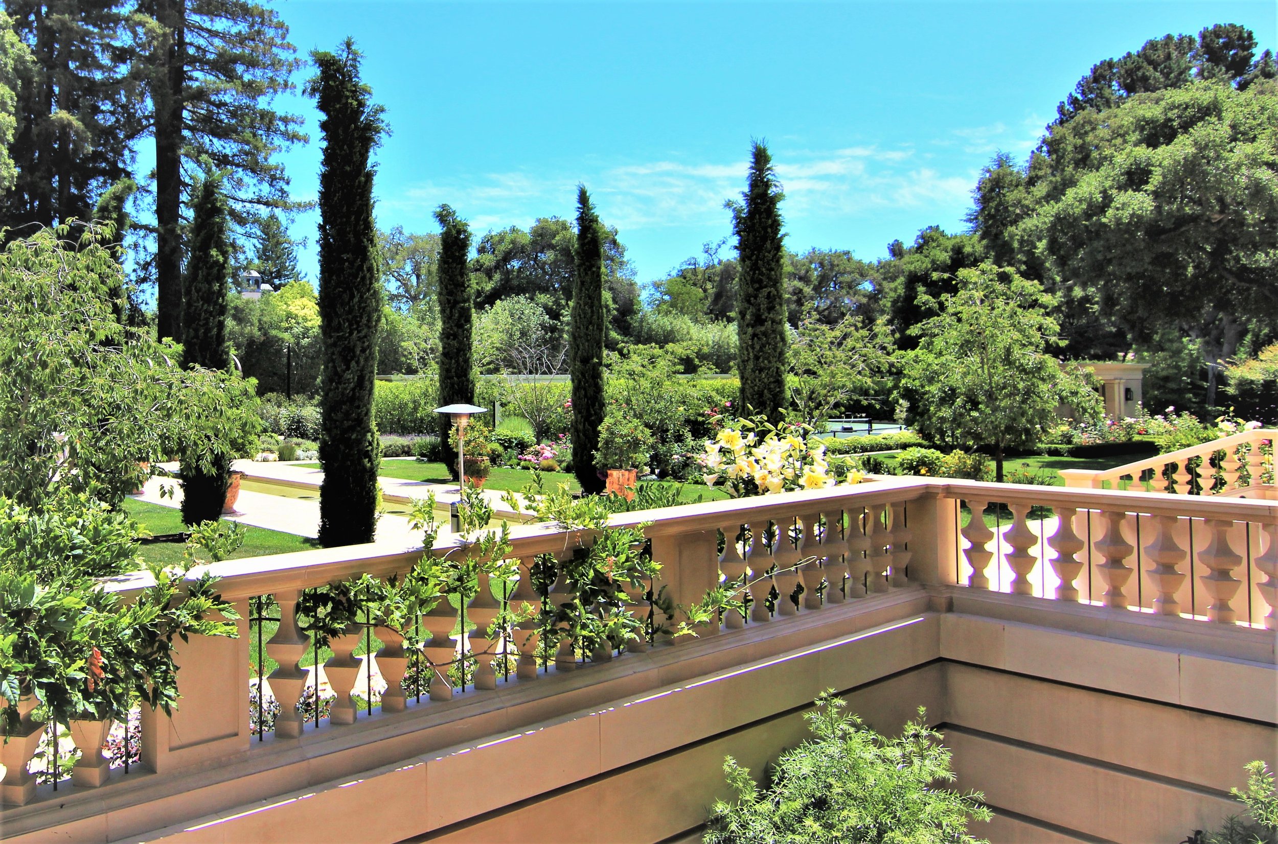 View from a balcony overlooking a garden with tall trees, trimmed shrubs, and a walkway, under a clear blue sky.