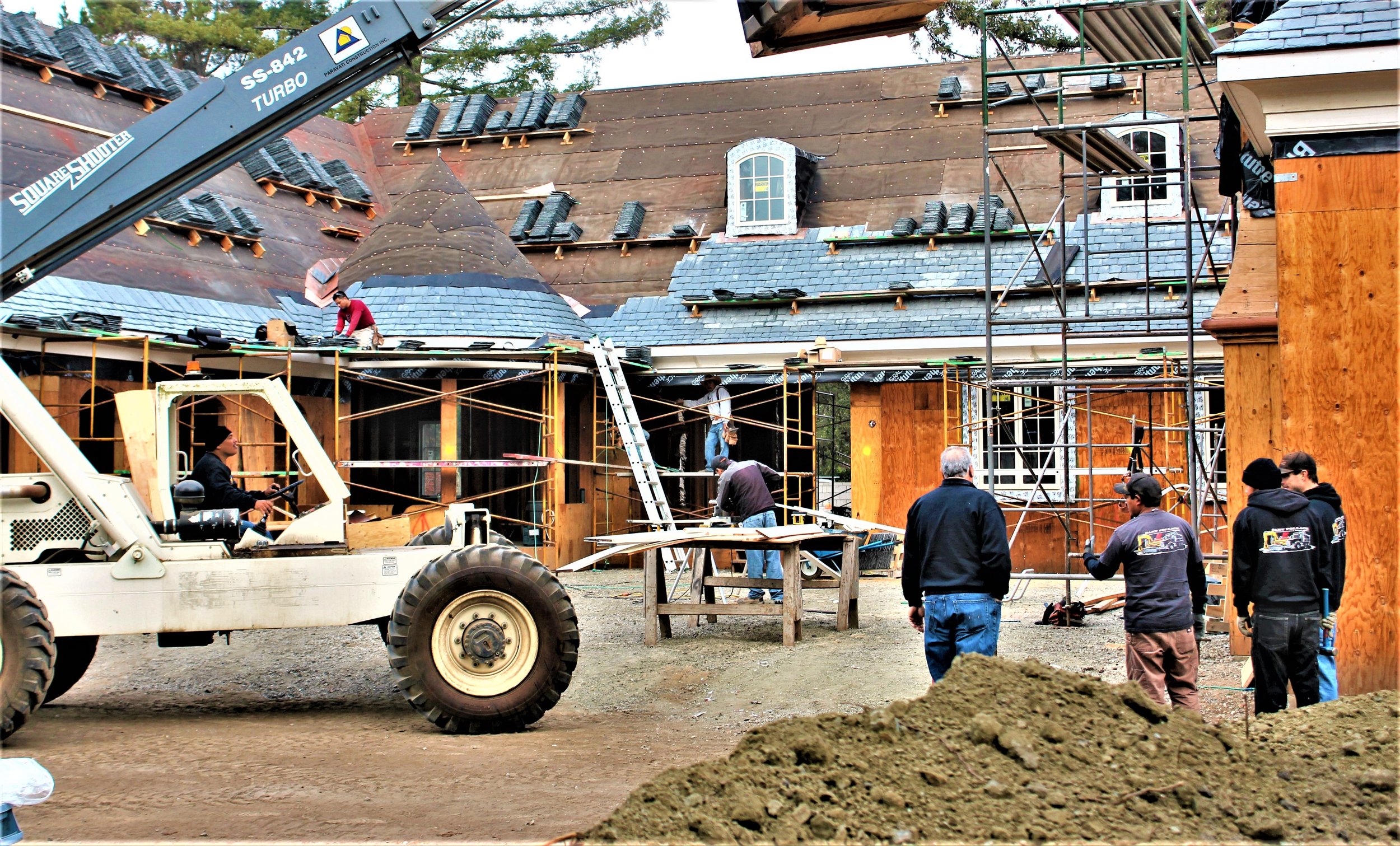 Construction workers building a house, scaffolding around the structure, a crane lifting materials, partially completed roof with shingles, and multiple people working on different sections.