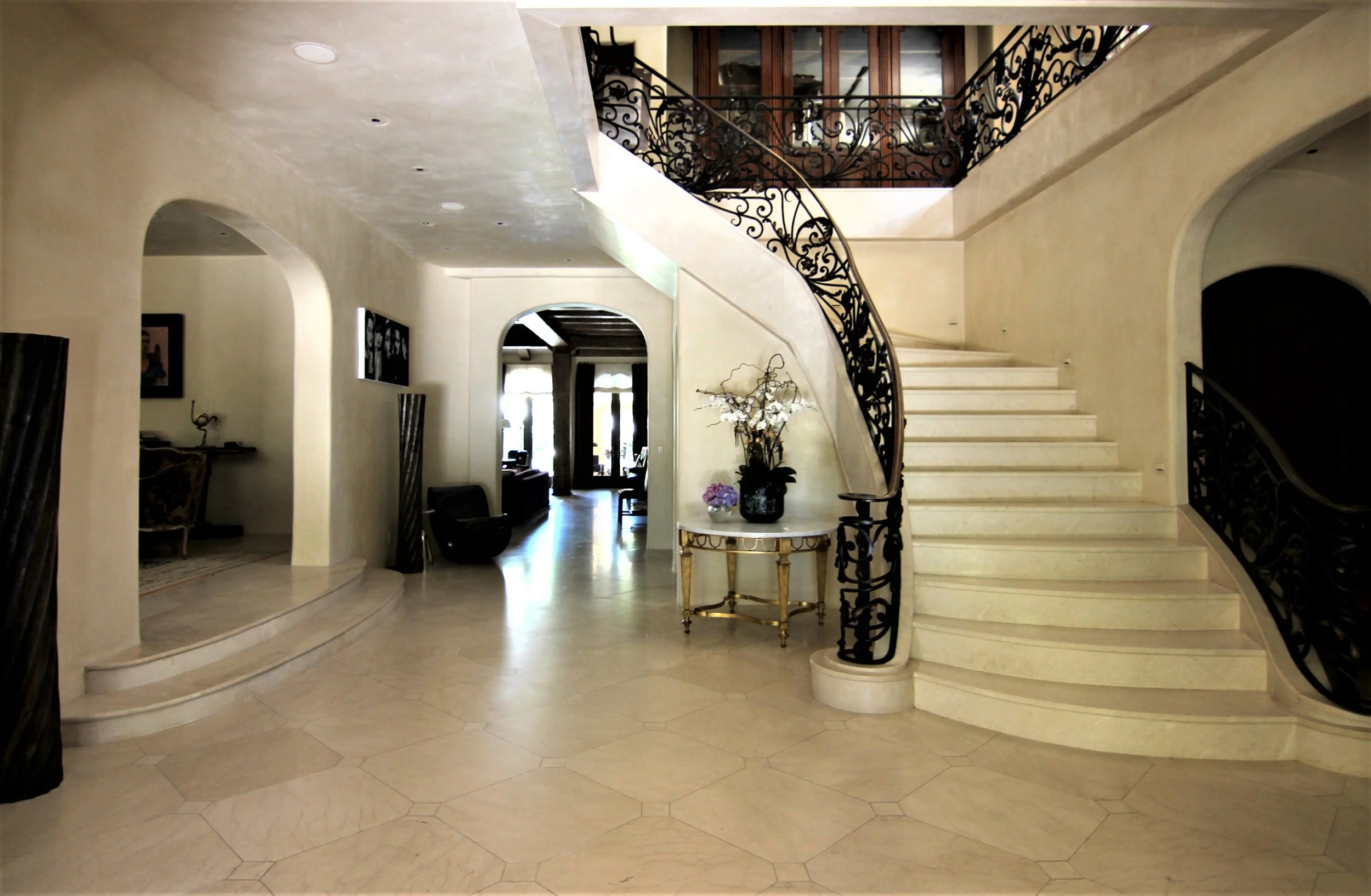 Interior view of a grand foyer with a curved staircase featuring ornate black wrought-iron railing, a small table with flowers, and multiple archways leading to other rooms.