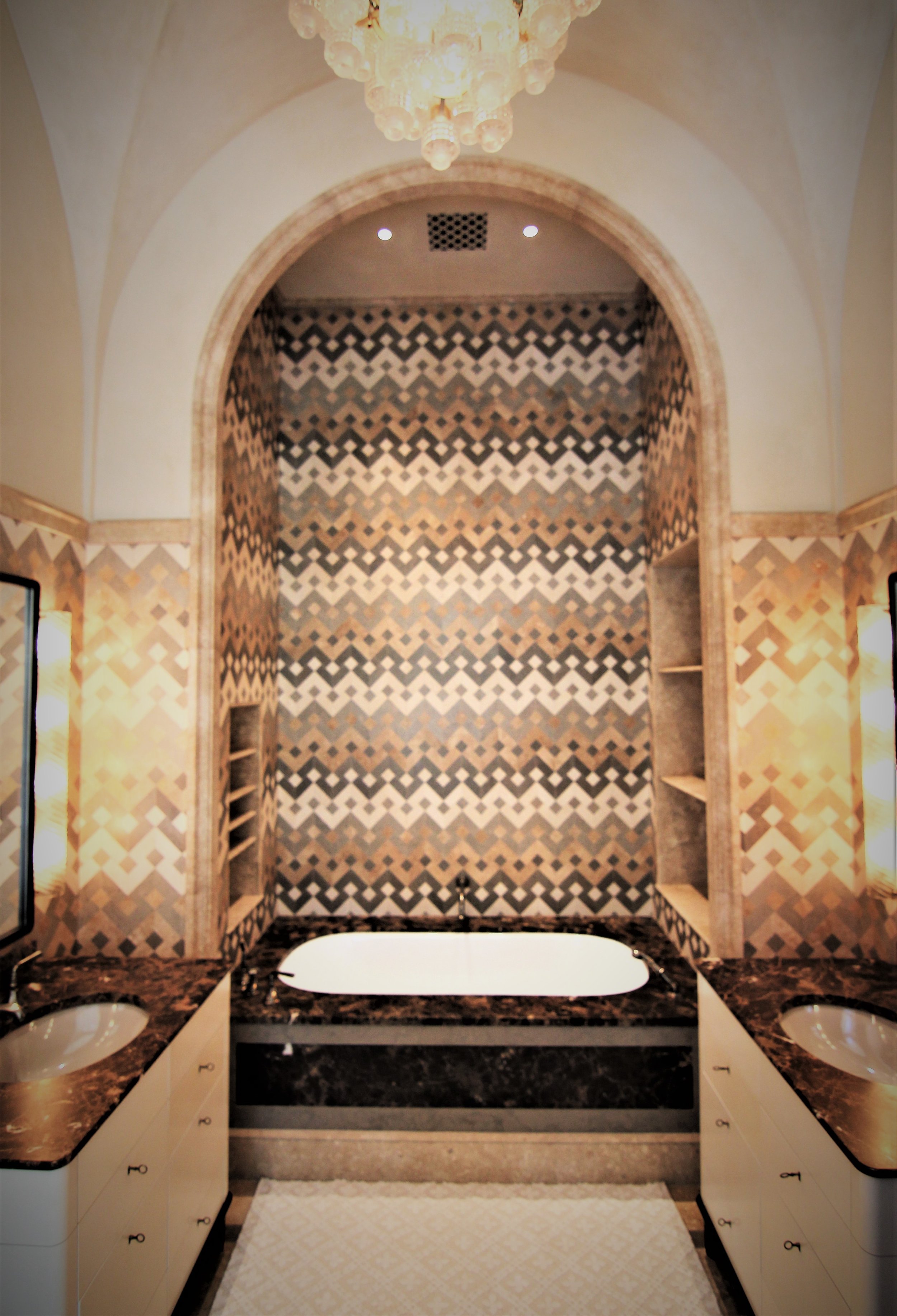 Luxurious bathroom featuring a double vanity with marble countertops, two sinks, a bathtub, and decorative tile walls with geometric patterns, illuminated by elegant lights and a chandelier.