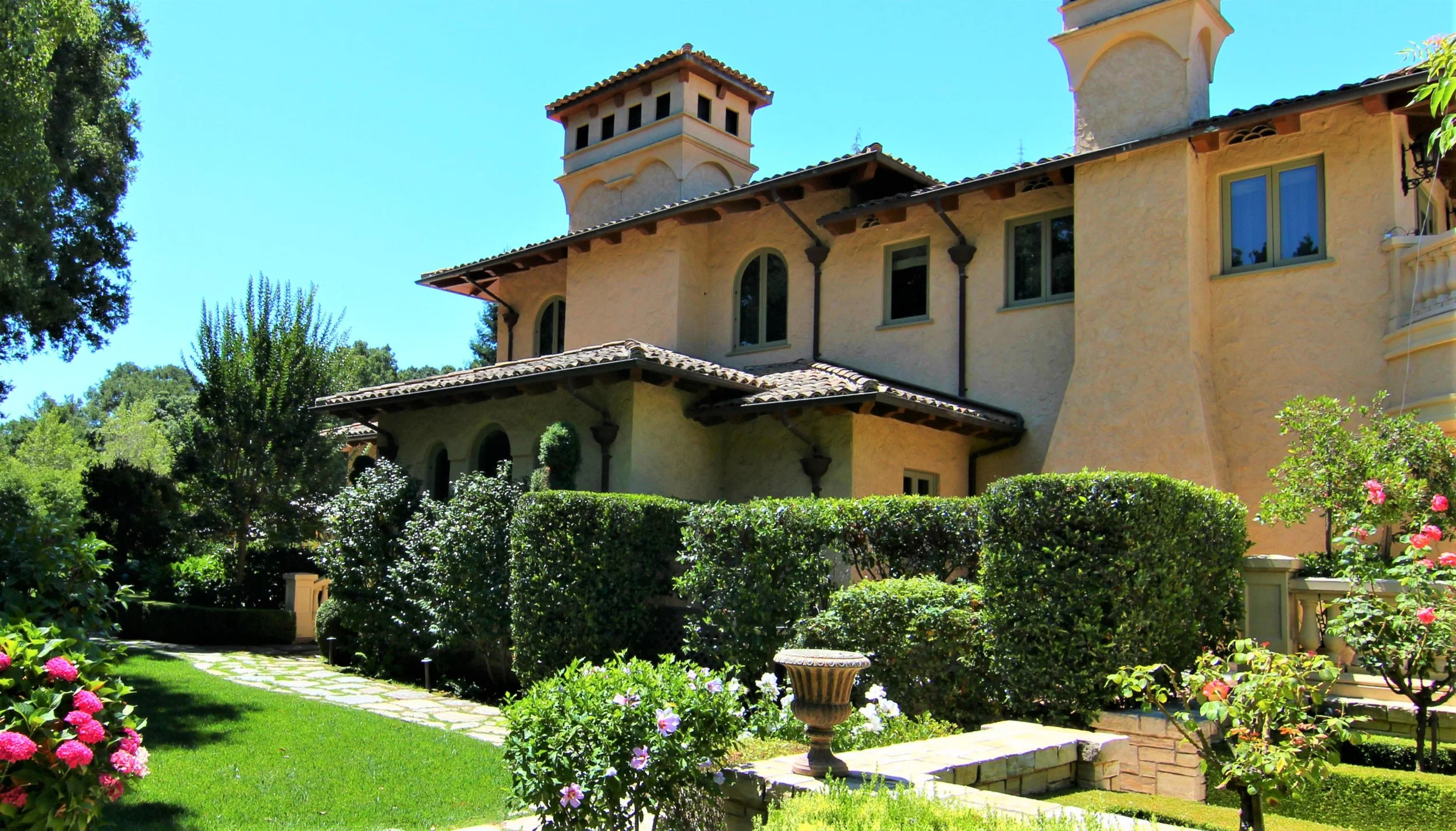 A large, elegant house with a stucco exterior and Spanish-style architecture, surrounded by a lush, well-maintained garden with trimmed hedges, colorful flowers, and green grass.