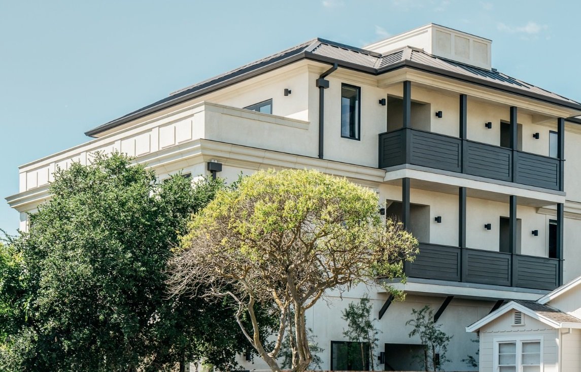 A modern, multi-story house with a flat roof, white exterior walls, and black balcony railings. There are trees and bushes in the front yard.