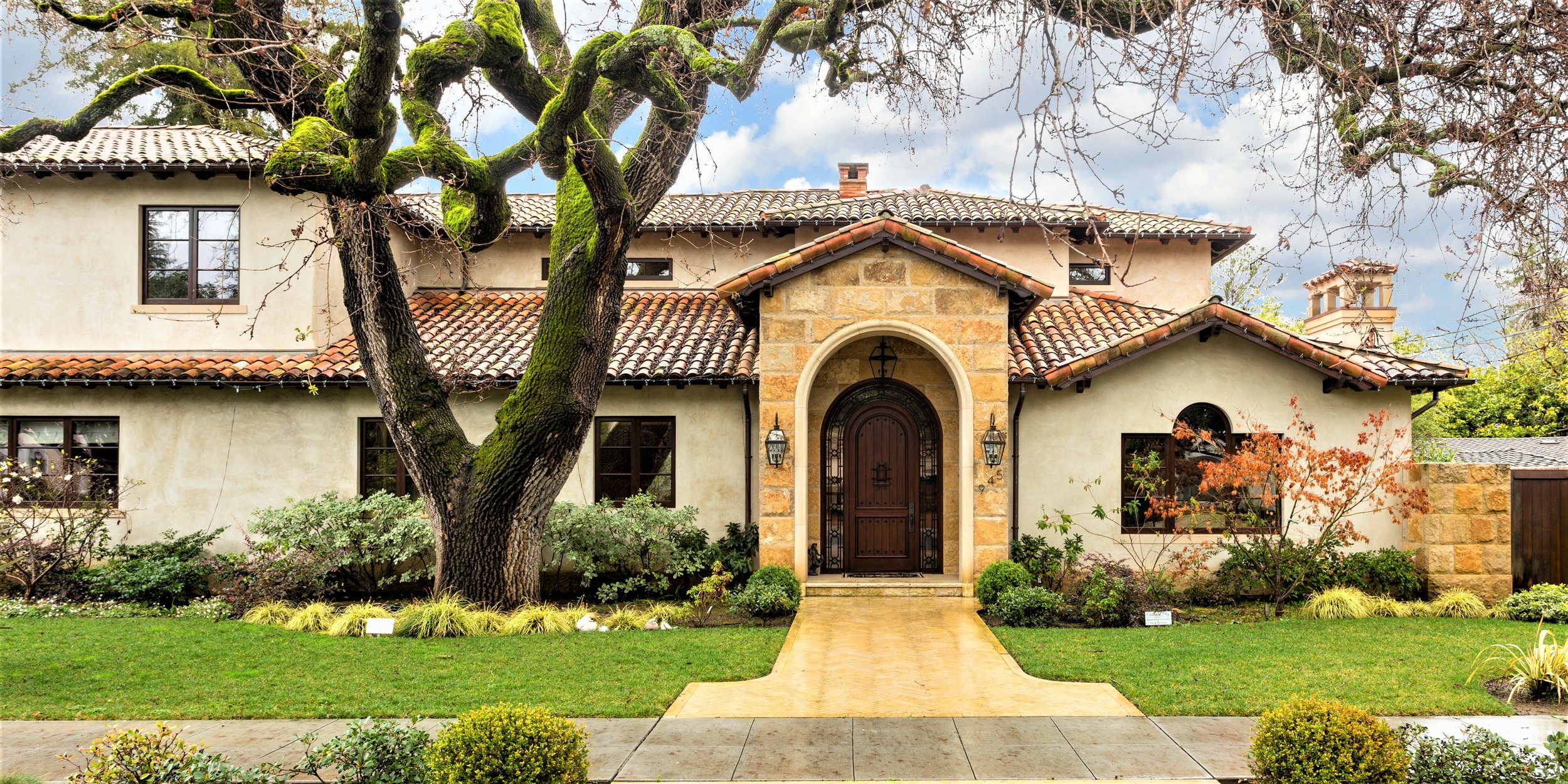 Front view of a house with a stone and stucco facade, red tile roof, and a large moss-covered tree in the front yard. The house has a wooden arched front door flanked by lantern-style light fixtures. There are well-maintained grass and shrubs along t
